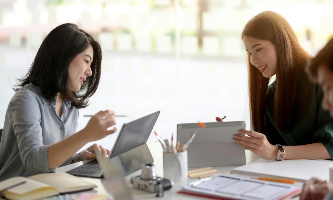 Two accounting professionals collaborating at a desk, one working on a laptop and the other on a tablet, surrounded by office supplies. Their interaction reflects effective capacity planning and teamwork.