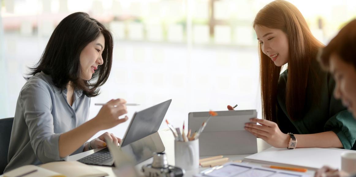Two accounting professionals collaborating at a desk, one working on a laptop and the other on a tablet, surrounded by office supplies. Their interaction reflects effective capacity planning and teamwork.