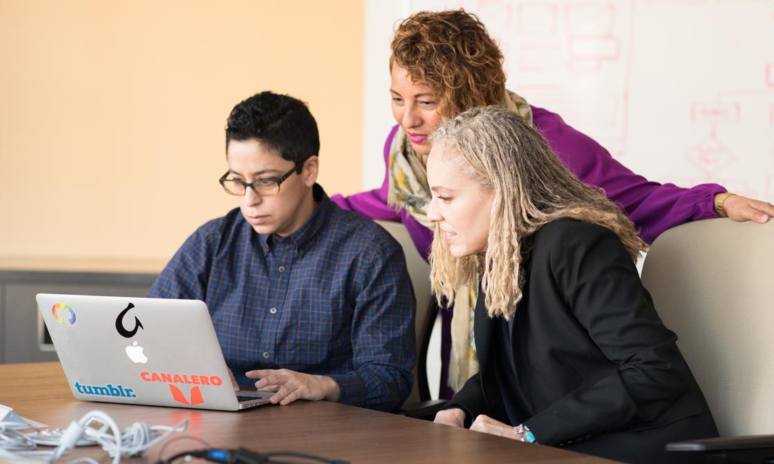 2 people are sitting at a desk with 1 person standing behind them. They're all looking intently at a laptop screen. There is a whiteboard with red writing on it behind them.