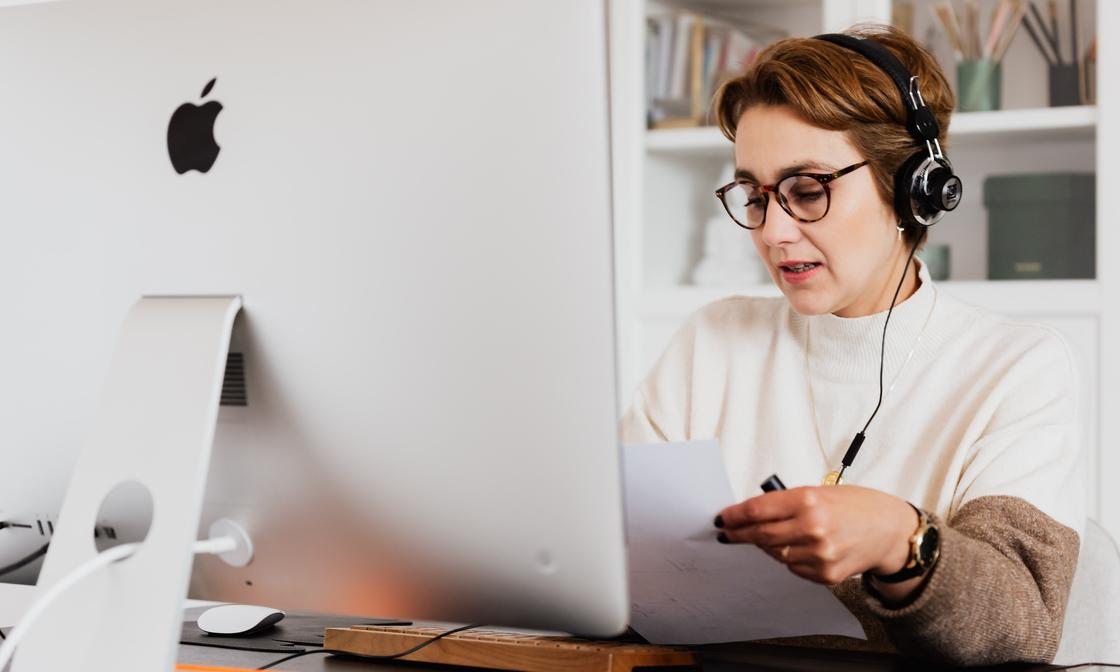 A person is working behind a desktop Mac. They're wearing a phone headset and they're holding a sheet of paper. It looks like they're on a call with a client.
