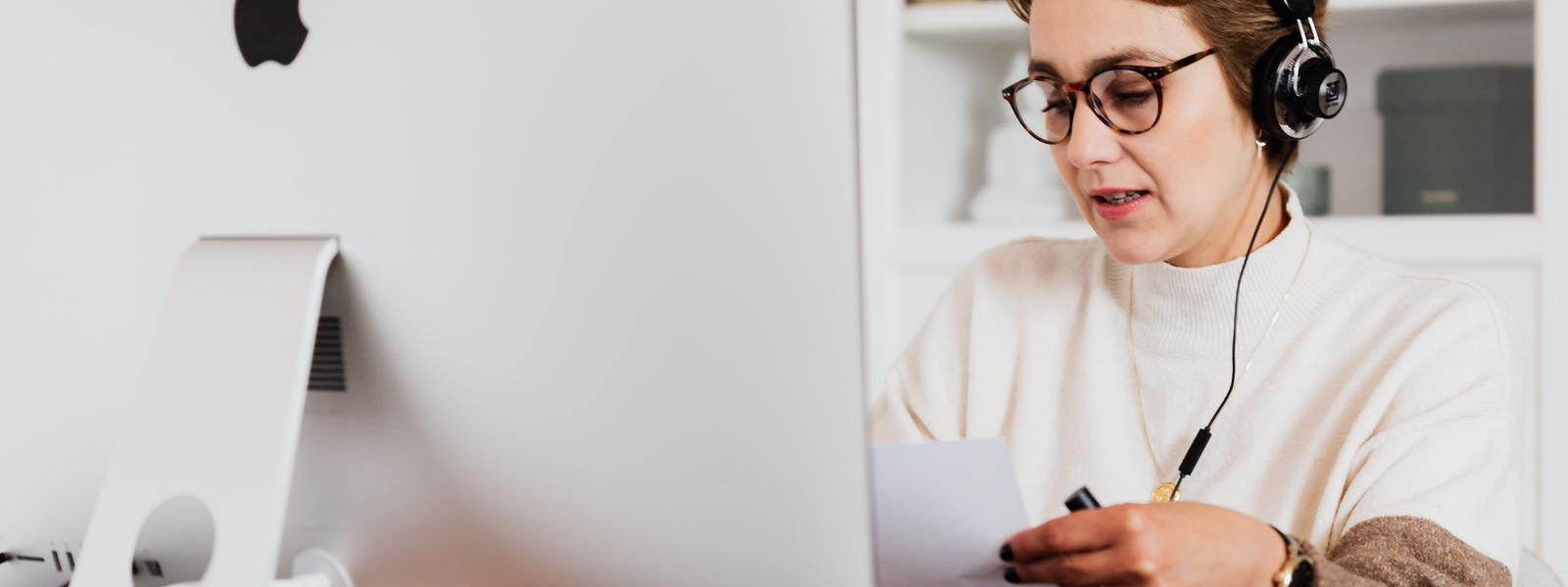 A person is working behind a desktop Mac. They're wearing a phone headset and they're holding a sheet of paper. It looks like they're on a call with a client.