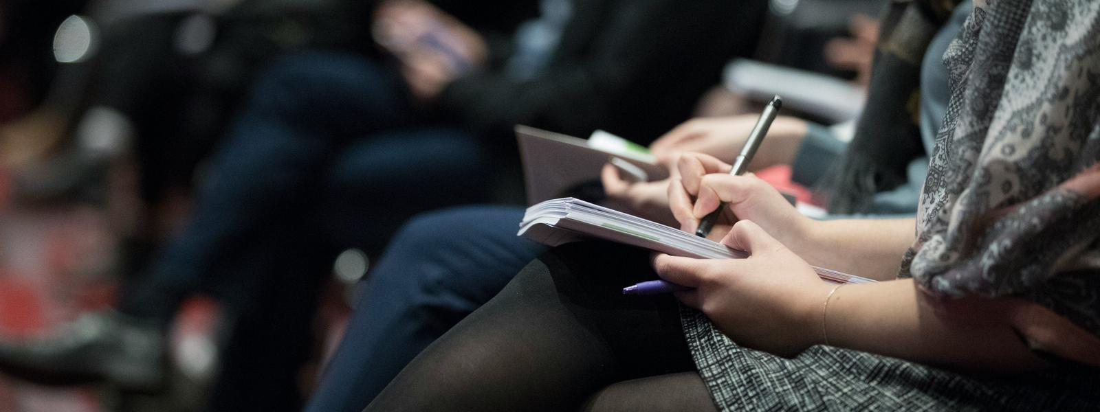 A row of hands taking notes in a room full of other accounting professionals.
