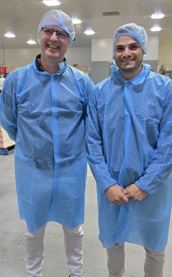 Two smiling men wearing blue disposable gowns and hairnets in a clean facility.