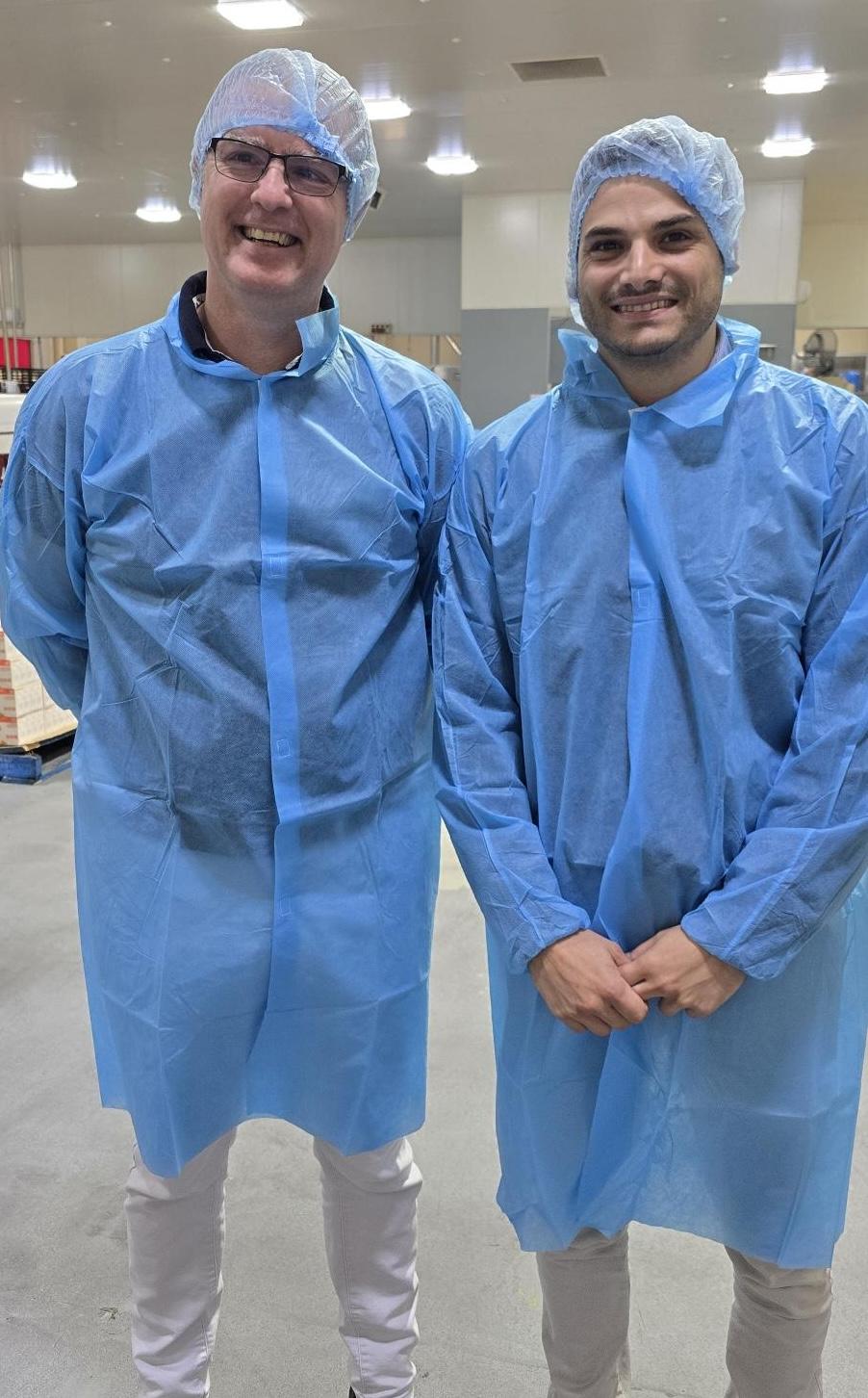 Two smiling men wearing blue disposable lab coats and hairnets stand in a clean production facility.