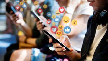 Close up of several teenagers looking at their phones, social media reactions like the thumbs up or heart are placed over the phone, suggesting that the teens are on social media