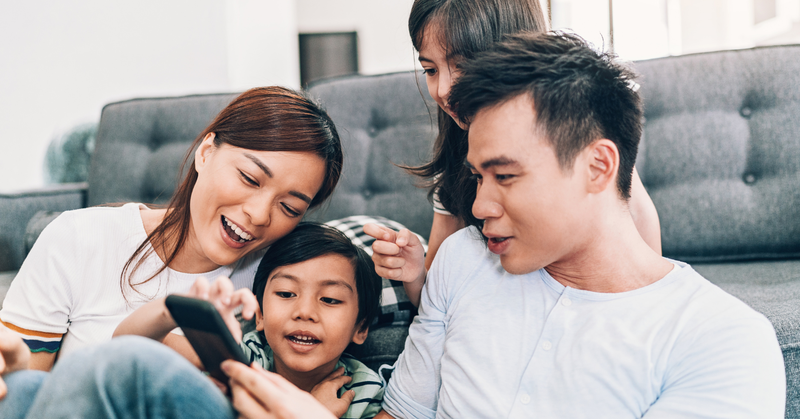 A family of four sits on a couch, smiling and looking at a smartphone together, creating a joyful, relaxed atmosphere.