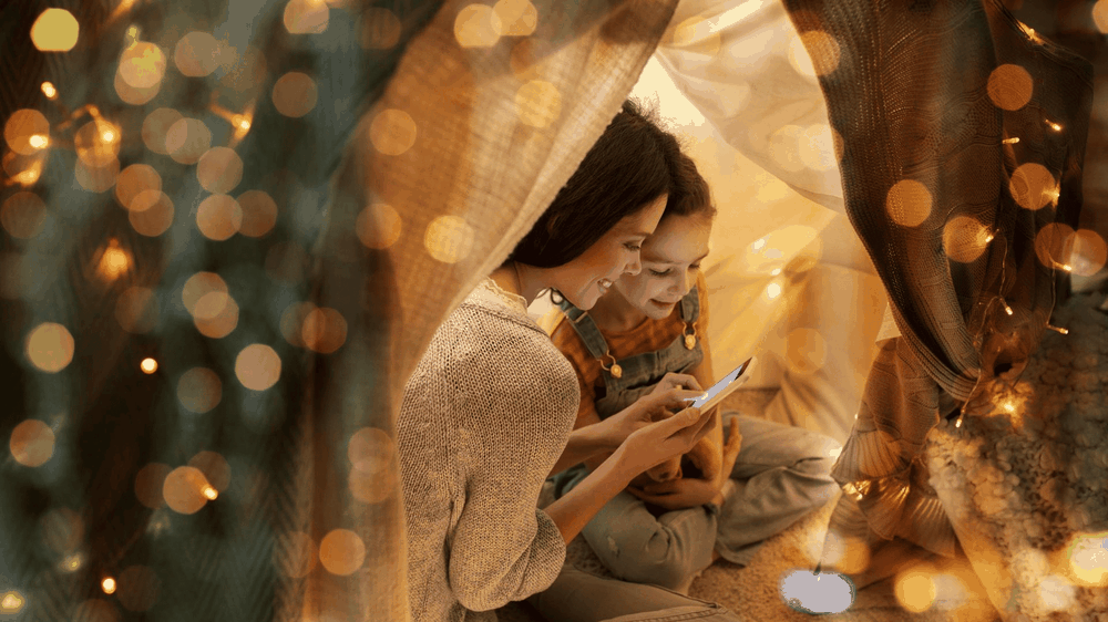 Woman and girl smiling inside a cozy blanket fort lit by fairy lights, looking at a glowing tablet.