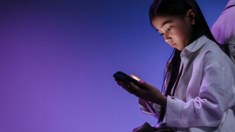 Young girl focused on a smartphone screen, surrounded by a purple glow, wearing a white jacket.