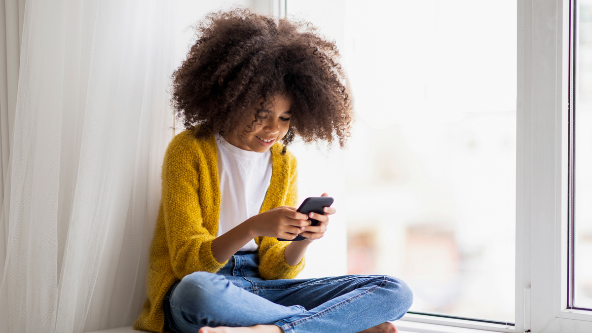 A young girl with curly hair and a yellow sweater is sitting by a window, looking at a smartphone with a smile.