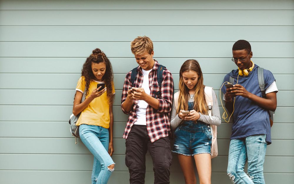 Four teenagers, two girls and two boys, lean against a wall and all of them are looking at their phones