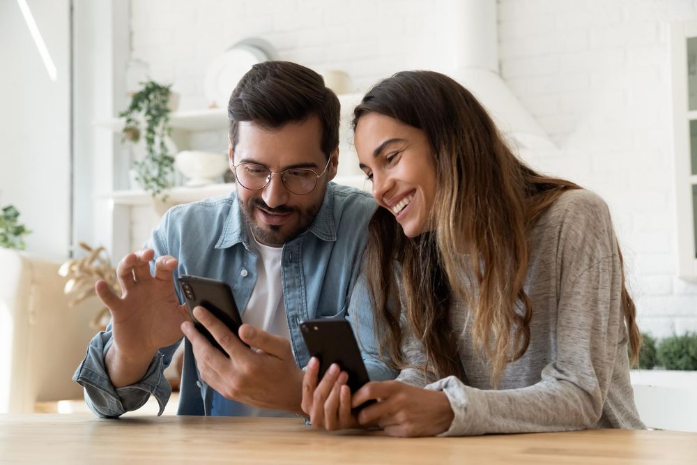 Parents sitting at table, both holding their phones in their hands, husband showing his wife something on his phone, she smiles