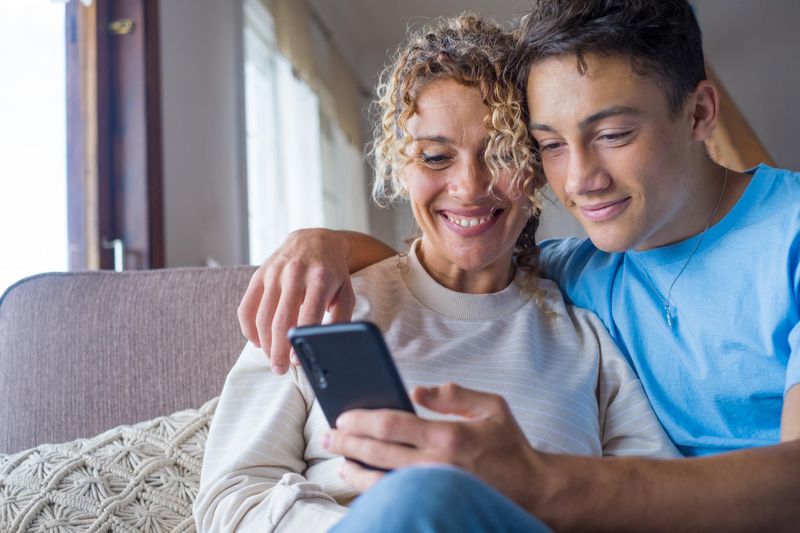 Smiling middle-aged 40s mother rest with teenage son using smartphone together, happy young man enjoy family weekend with mom taking a selfie, have fun at home