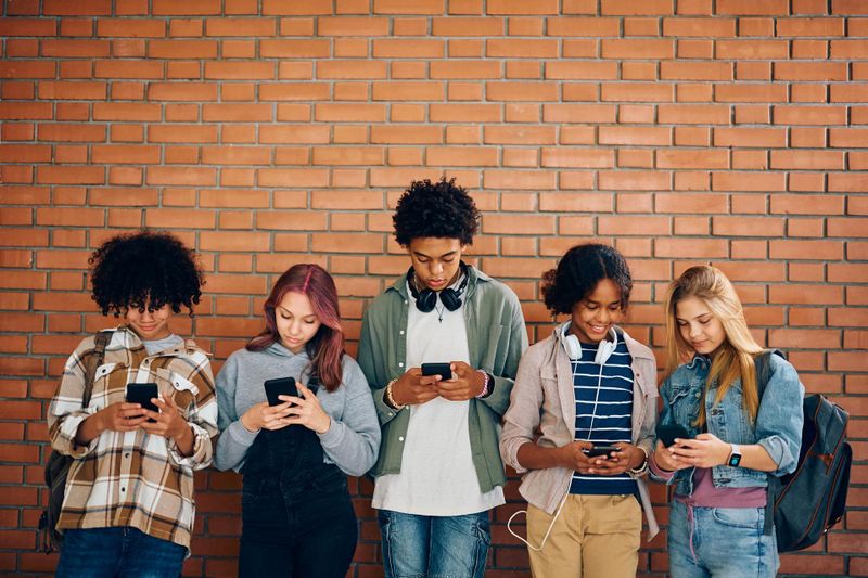 Multiracial group of teenagers using their cell phones while standing against the wall