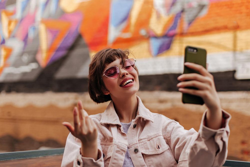 Teenager with pink glasses takes selfie in front of graffiti sprayed wall