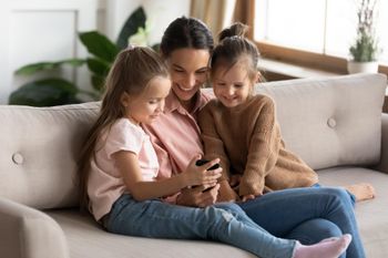 Mom is sitting on the couch with her two little girls, they all smile while looking at a smartphone screen