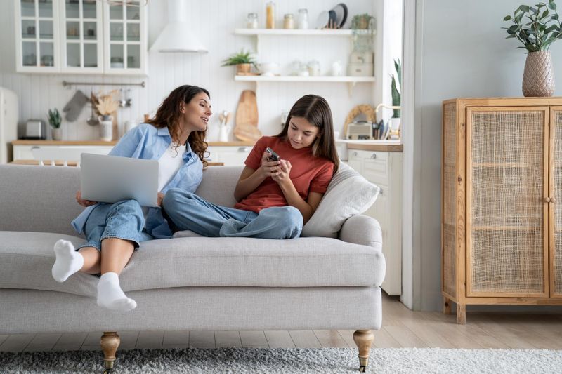 Mother and daughter are sitting on the couch, mom is trying to see what her daughter is doing on her smartphone