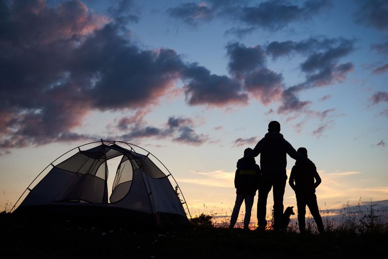 Rear view of silhouette of a father backpacker hugging two children. Tourists guys and dog stands beside camping on the top of mountain near tent at sunset under evening sky with clouds