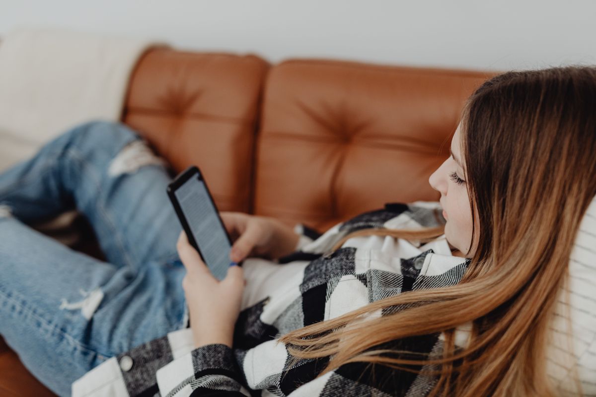 A teenage girl in a plaid shirt lies on a brown couch, focused on reading from a smartphone.