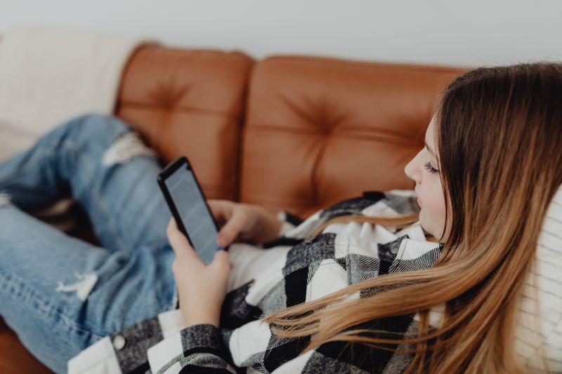 A teenage girl in a plaid shirt lies on a brown couch, focused on reading from a smartphone.