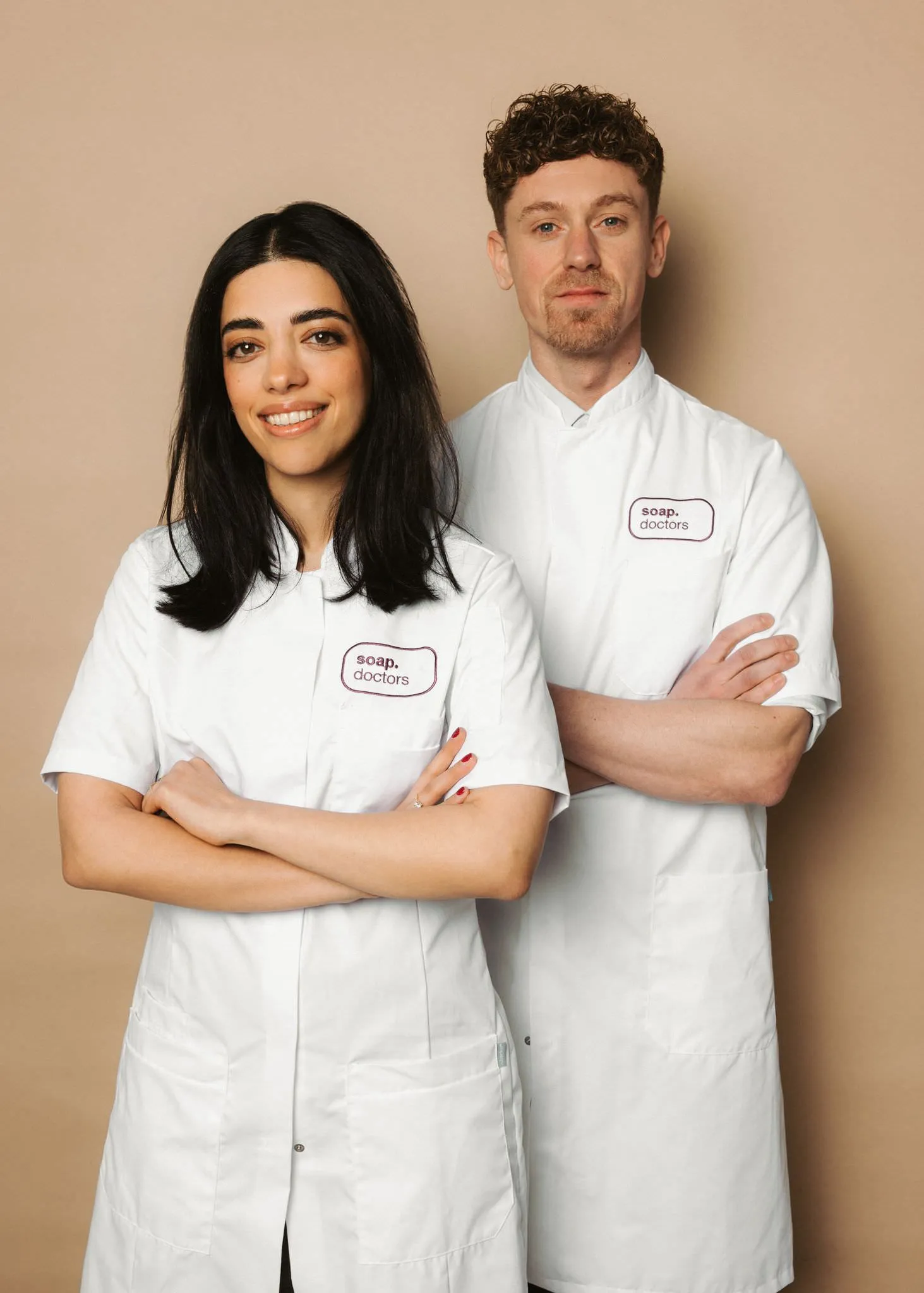 Two people in white lab coats labeled "Soap Doctors," standing confidently with arms crossed, smiling against a beige background.