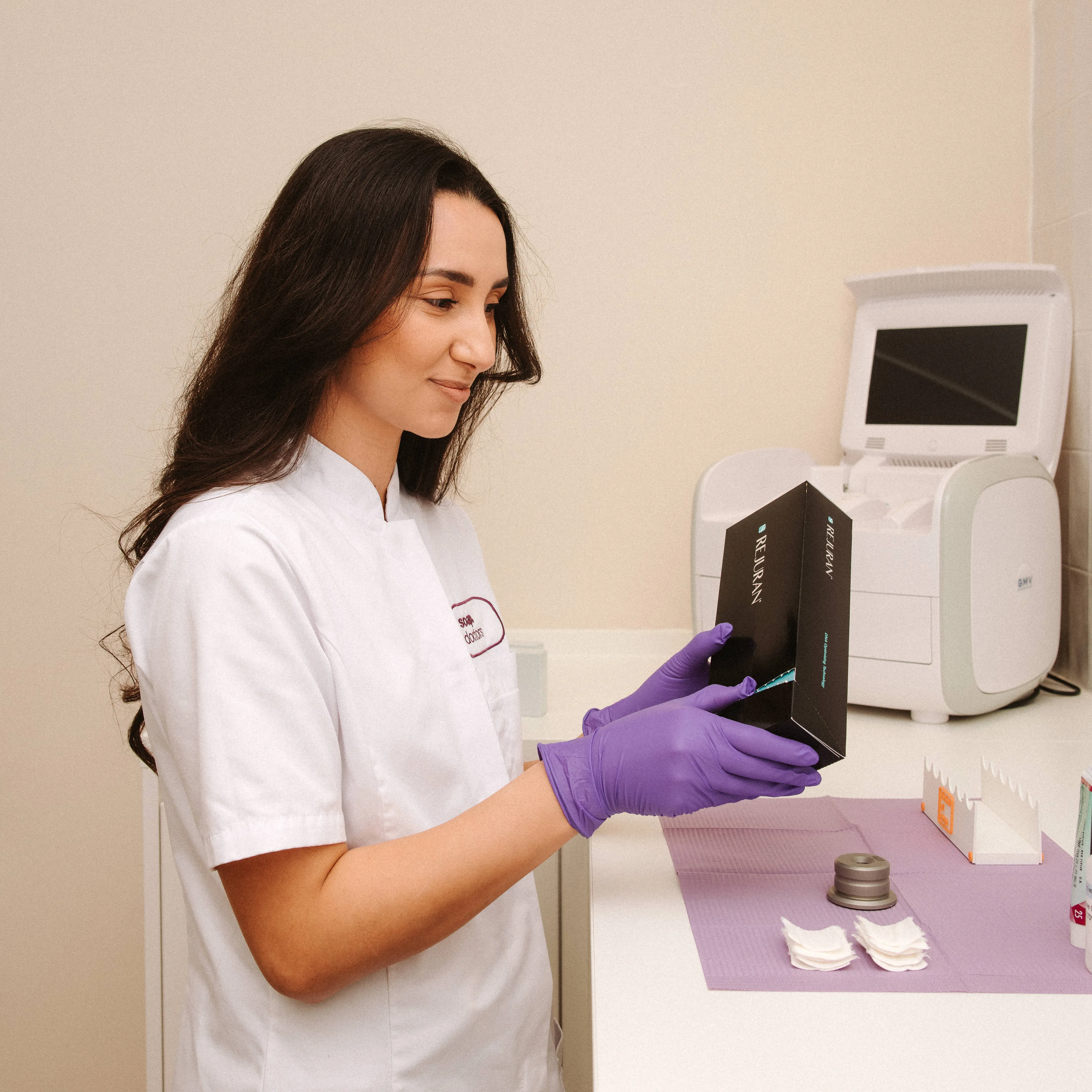 A woman in a white coat and purple gloves holds a black box in a lab setting with equipment and supplies on a counter.