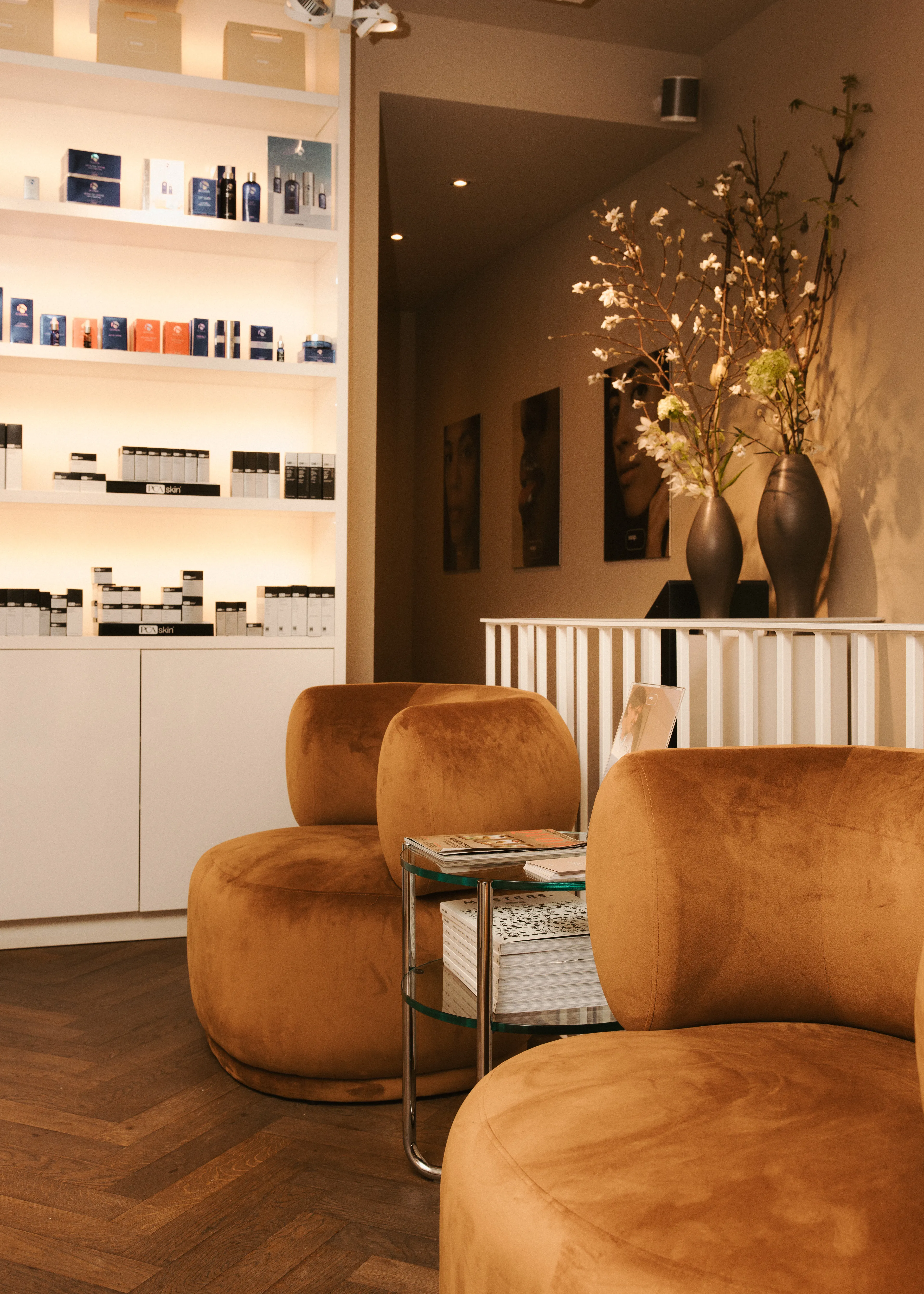 Cozy waiting area with brown velvet chairs, wooden flooring, a shelf with beauty products, and decorative vases with branches on a counter.
