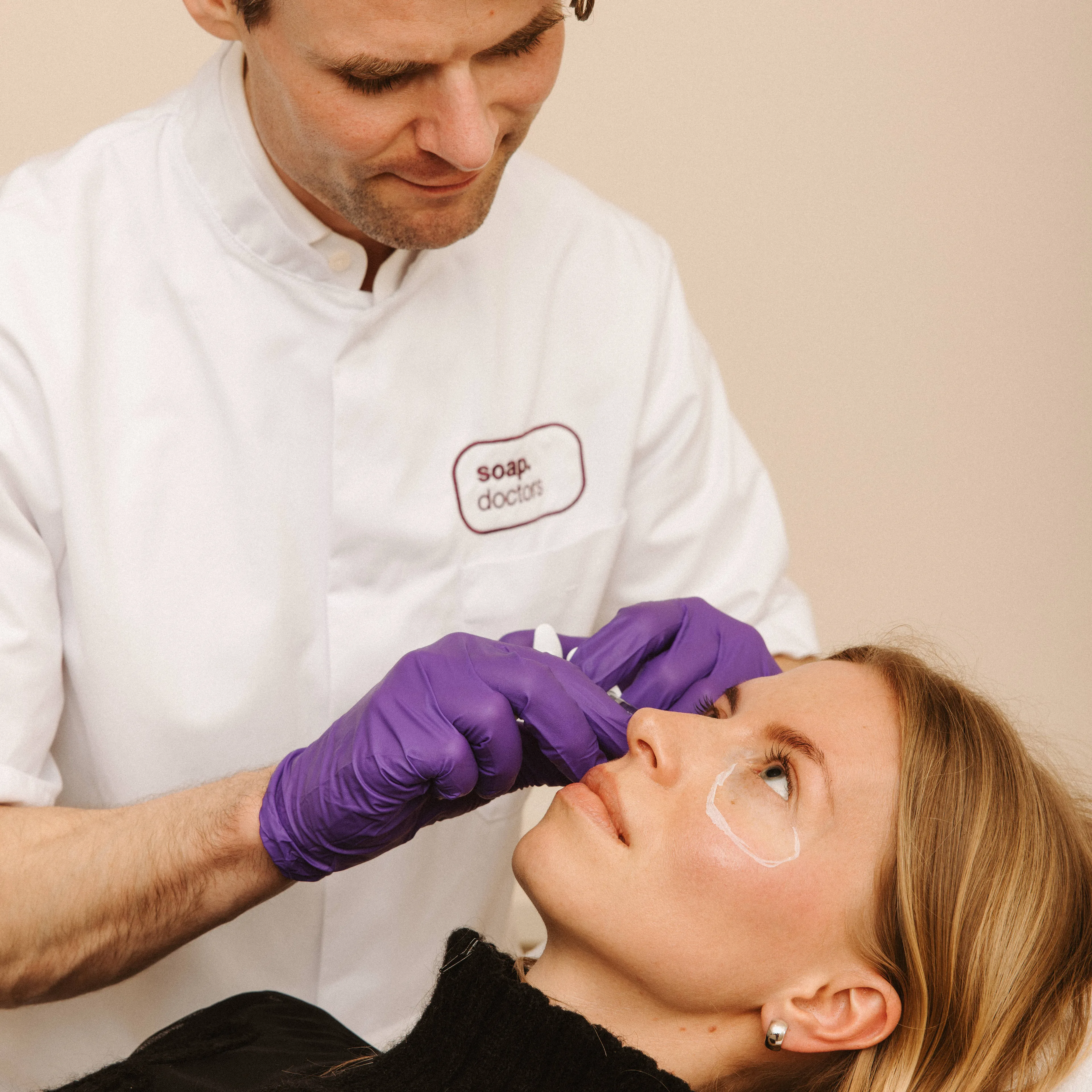 A professional in a white coat and purple gloves performs a cosmetic procedure on a woman lying down, with eye treatment patches applied.