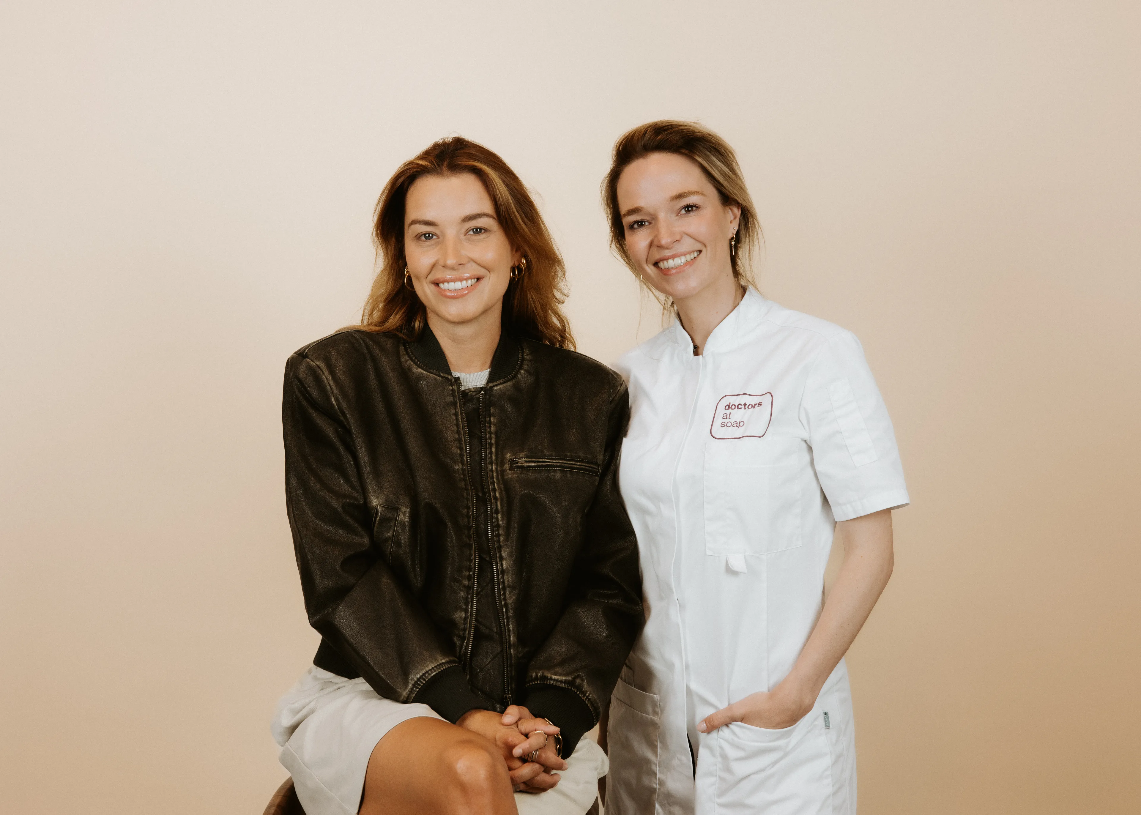 Two smiling women pose together; one in a black jacket sits, and the other in a white coat stands beside her against a beige background.
