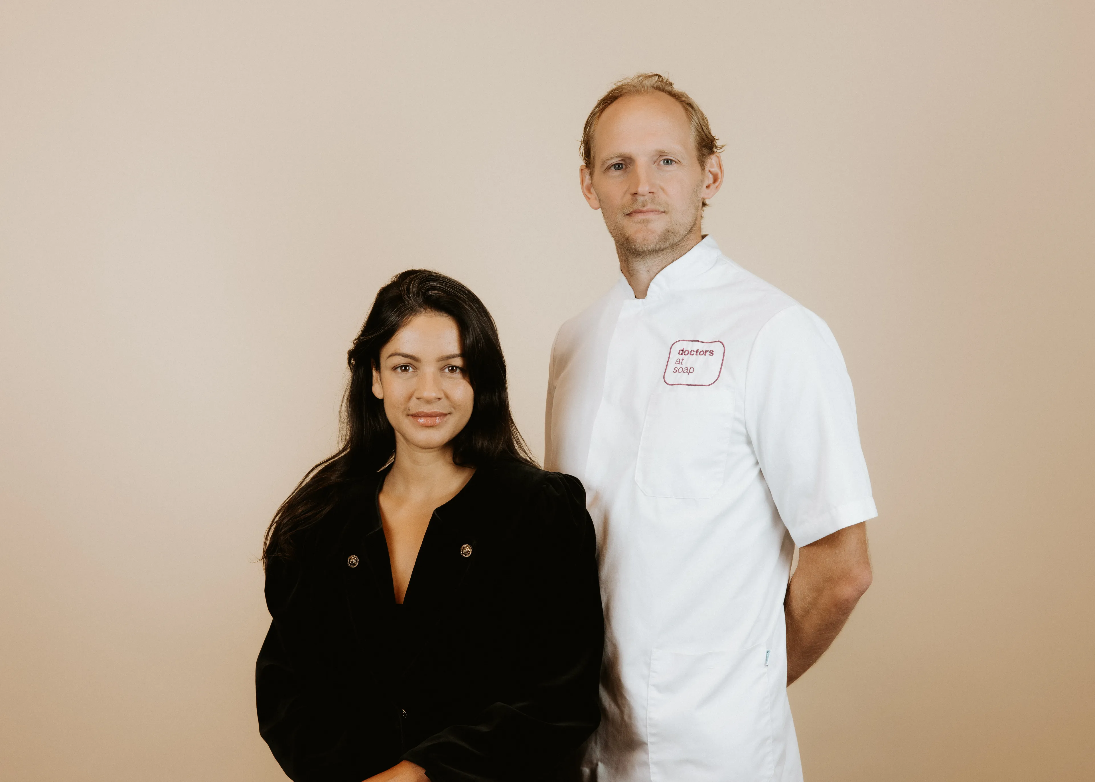 A woman in a black blouse and a man in a white chef's coat stand side by side against a plain background.