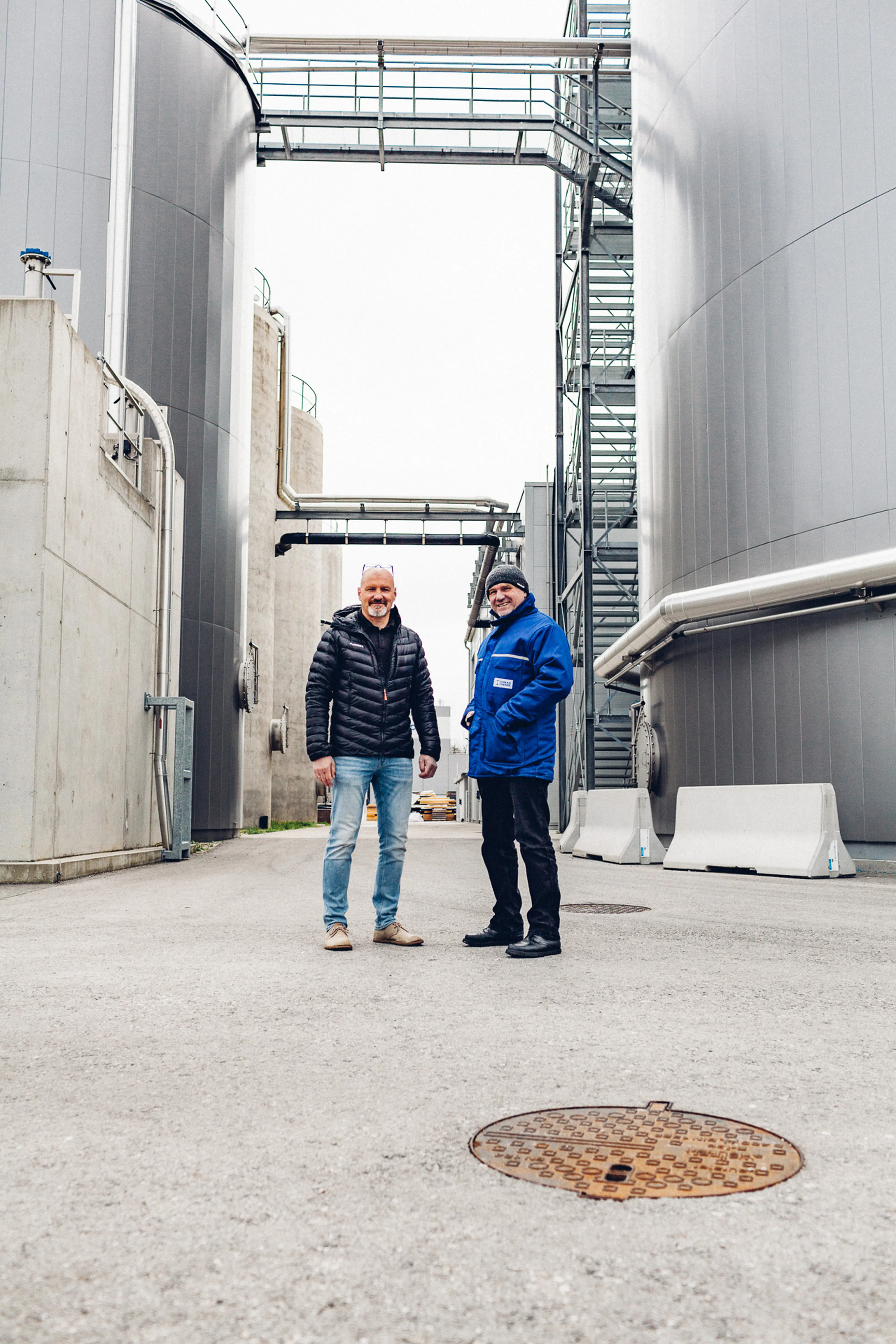 Two men stand smiling in an industrial setting between large metal tanks, wearing casual jackets and jeans. Pipes and structures are visible above.