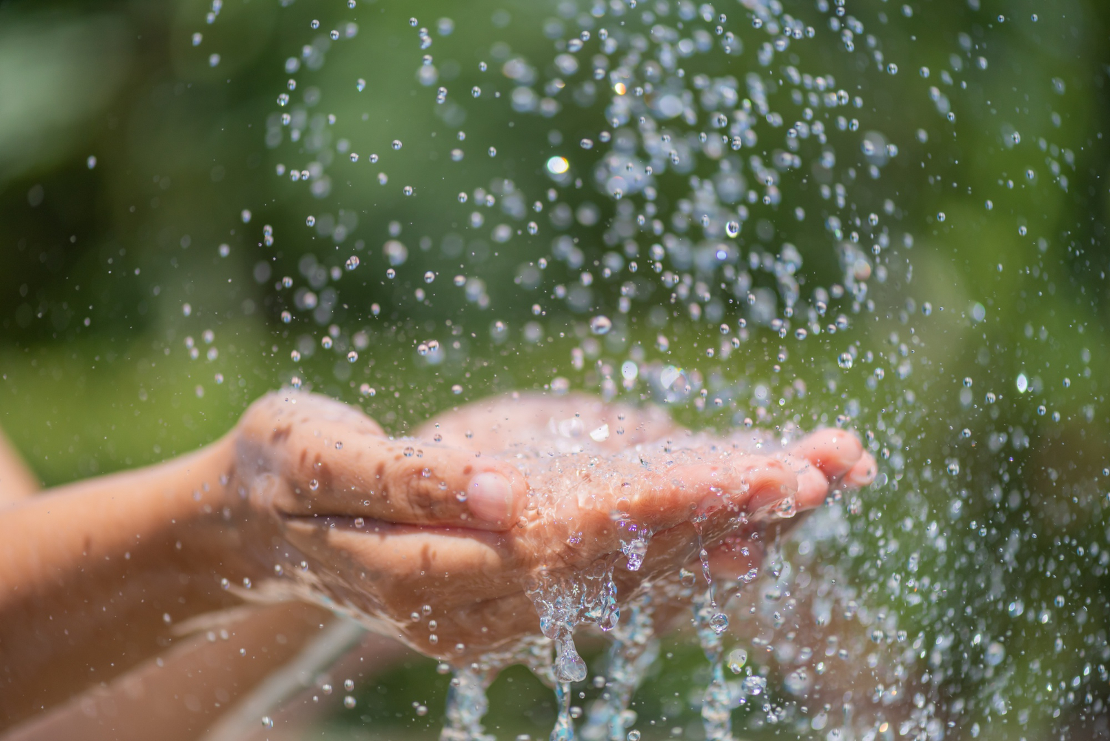 Hands cupped together catching and splashing clear water droplets, with a blurred green background.