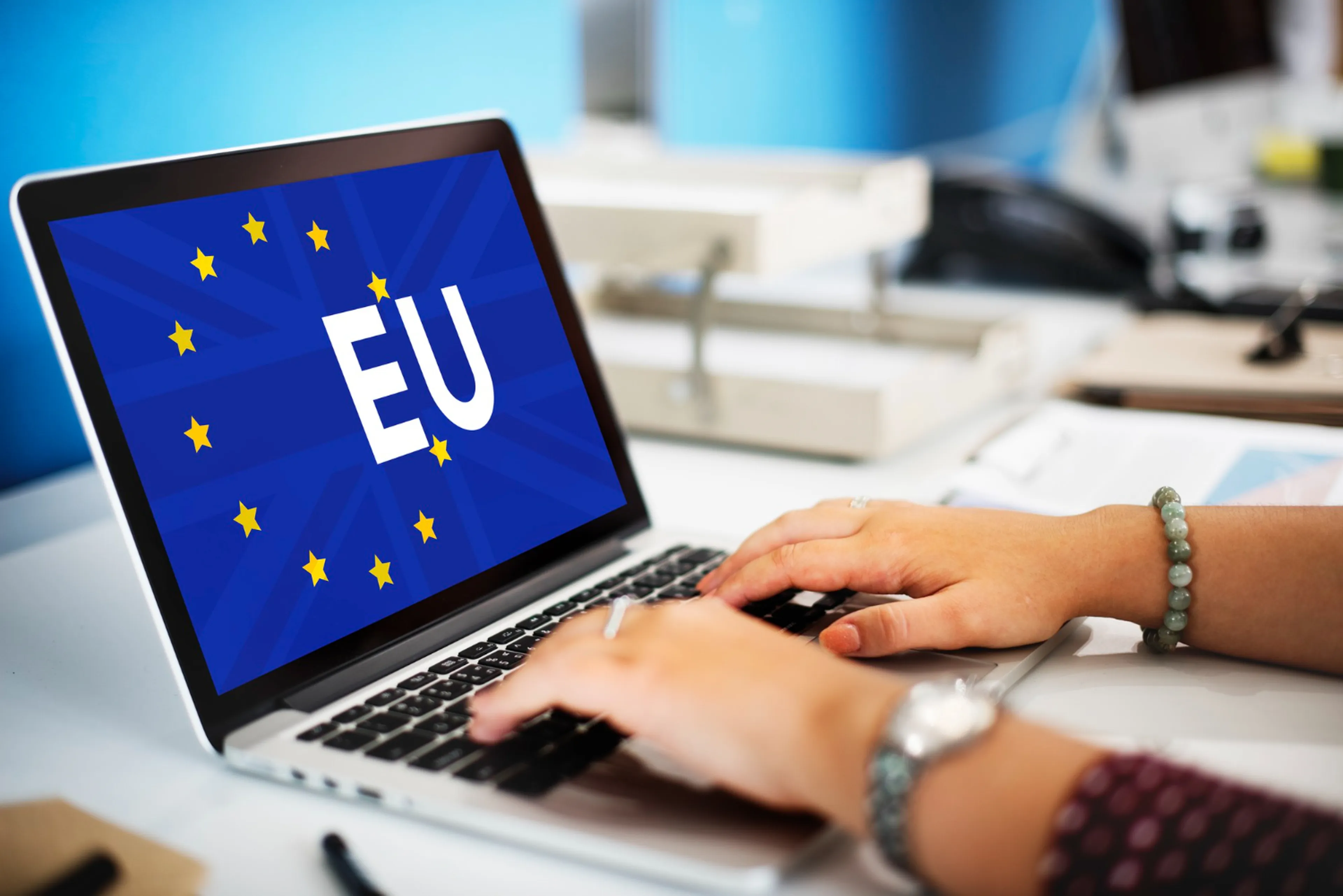 Hands typing on a laptop with an EU flag on the screen, featuring yellow stars in a circle against a blue background.