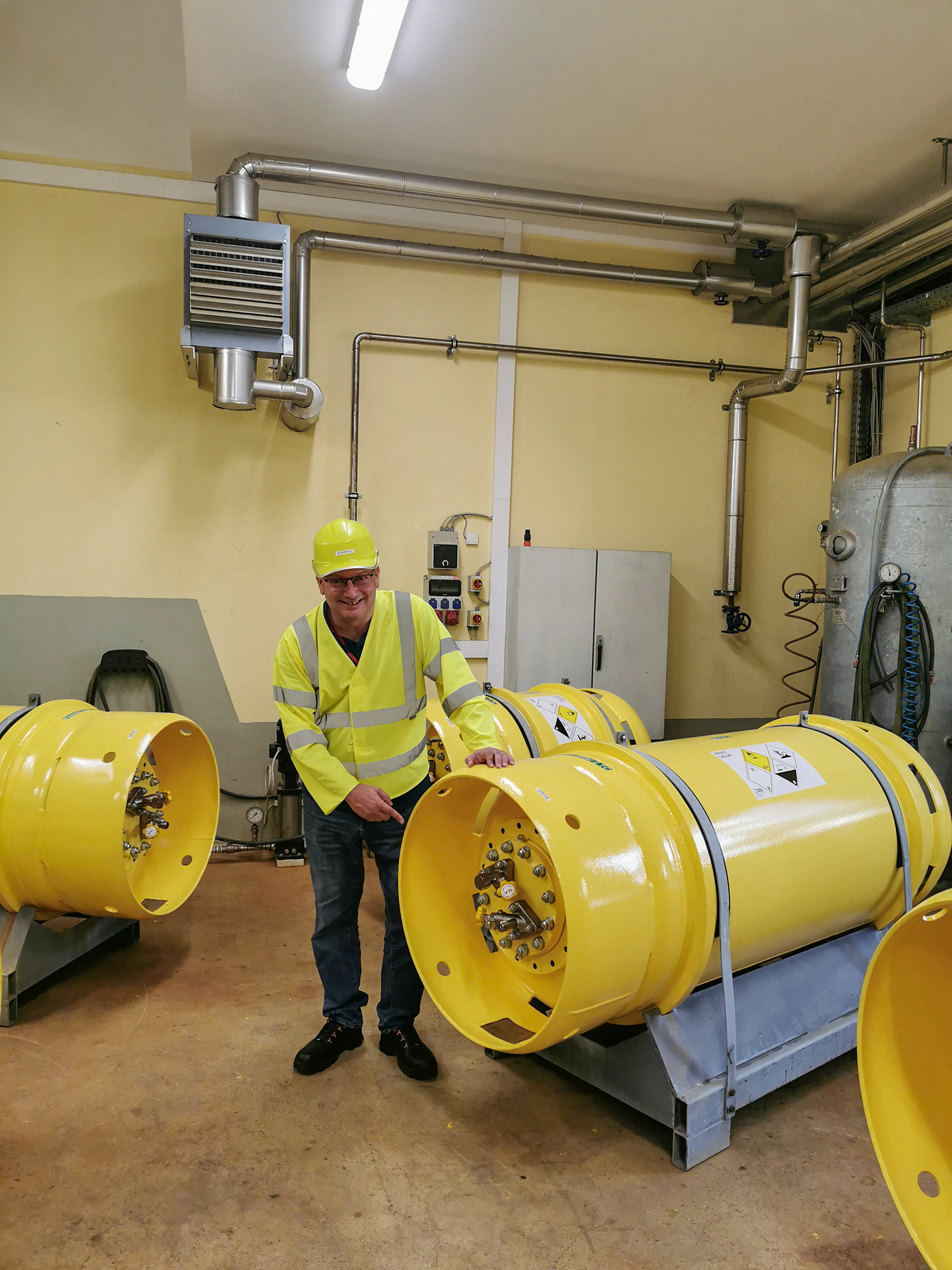 A person in a yellow safety helmet and vest stands in an industrial room, next to large yellow machinery parts. Pipes and equipment are visible.