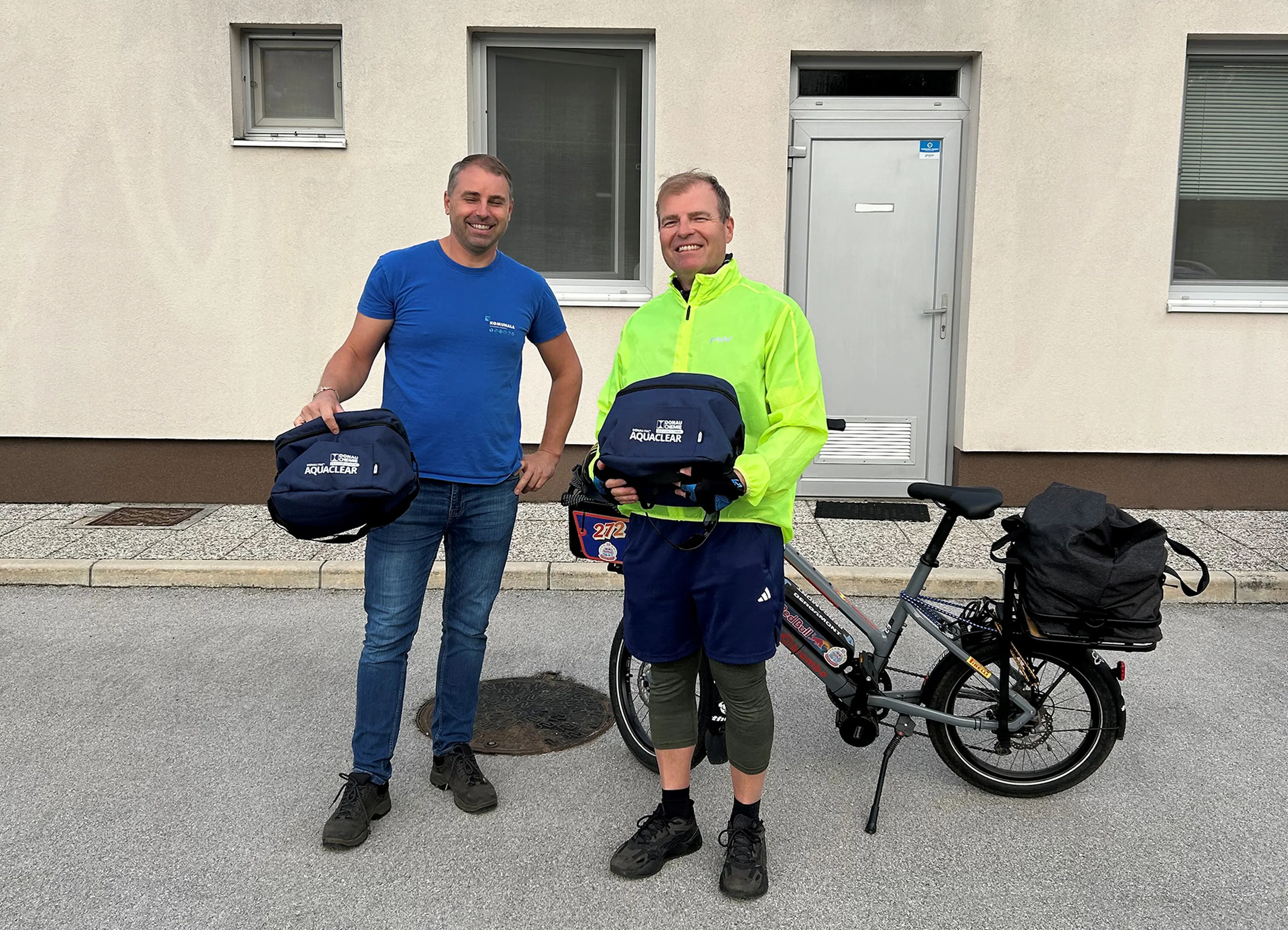 zwei Personen mit Taschen in der Hand auf denen das Logo der Donau Chemie Wassertechnik drauf ist, Fahrrad im Hintergrund