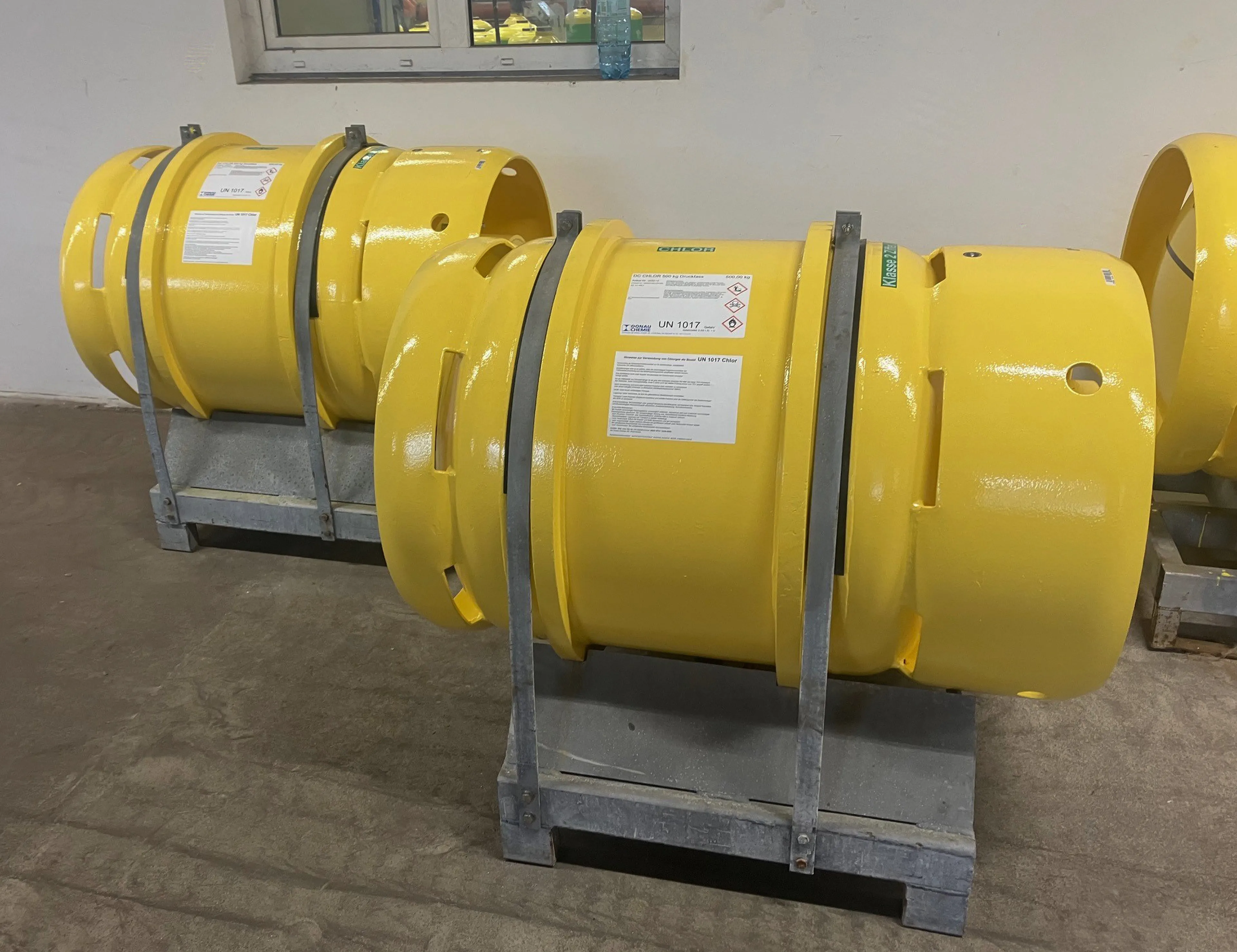 Two large yellow industrial cylinders with labels, secured on metal frames, placed on a concrete floor in a room with a small window.