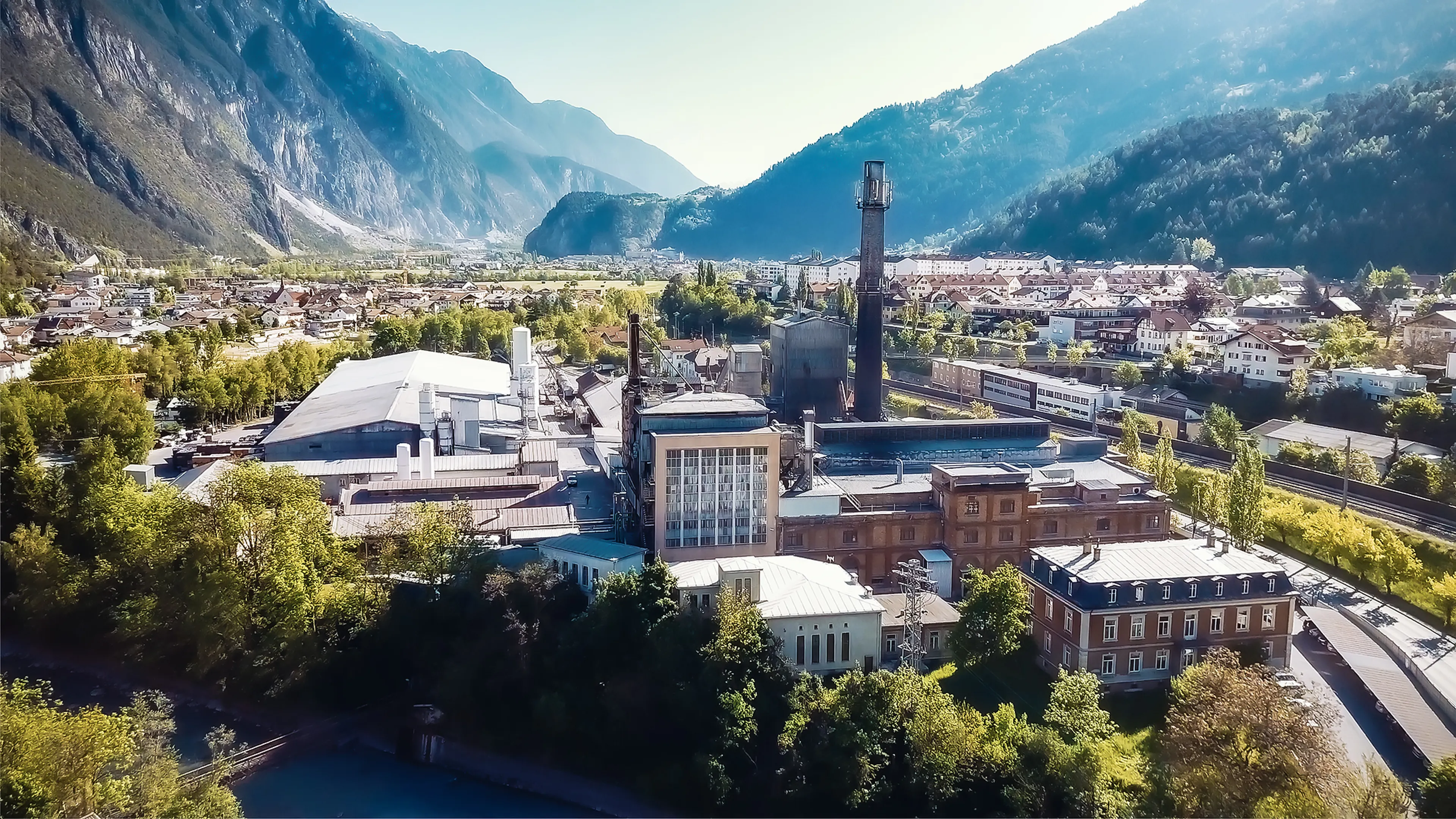 Aerial view of an industrial complex surrounded by trees and mountains, with a town and railway track nearby under a clear sky.