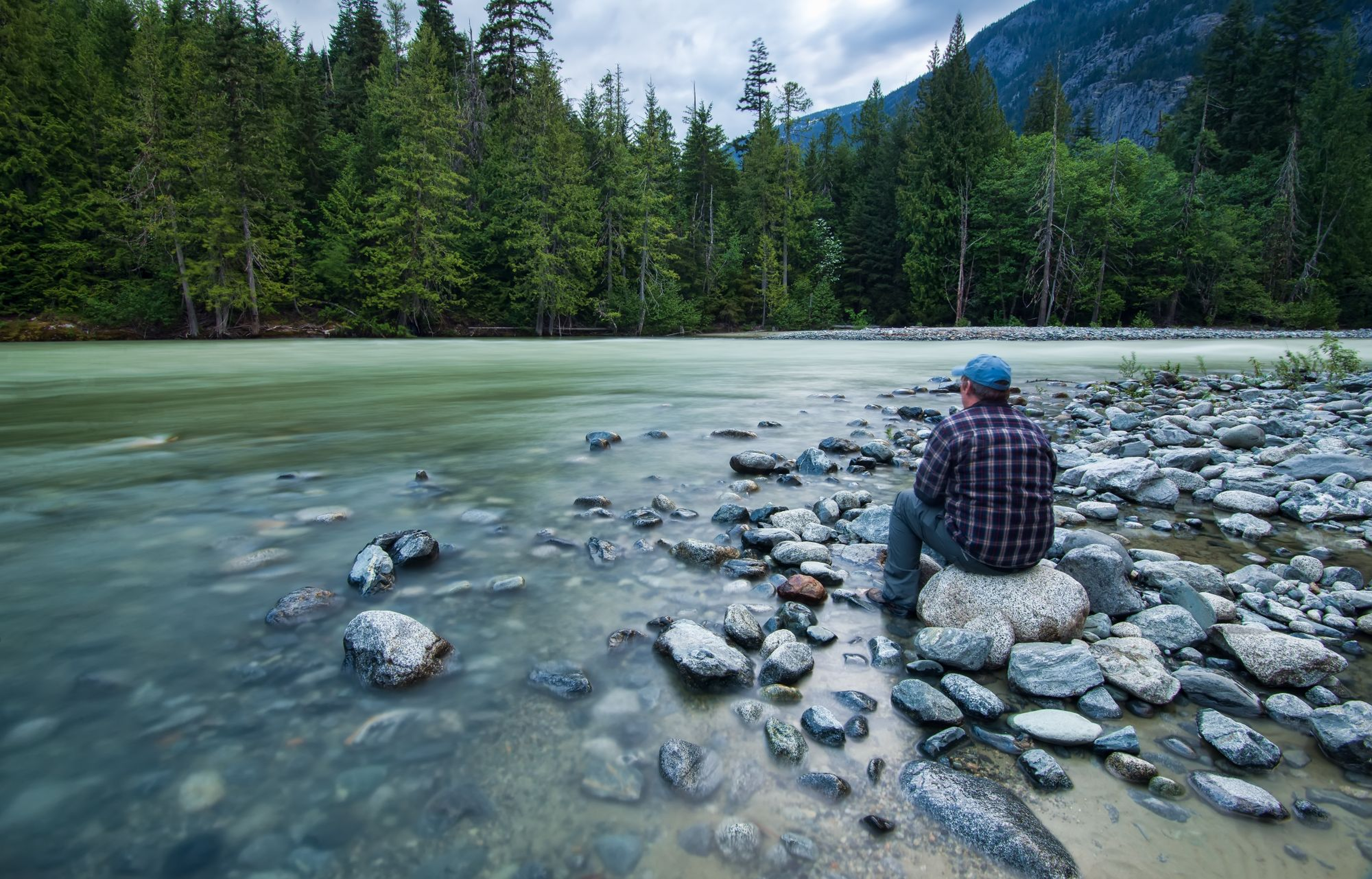 Person sitting on rocks by a gently flowing river, surrounded by forest and mountains under a cloudy sky.
