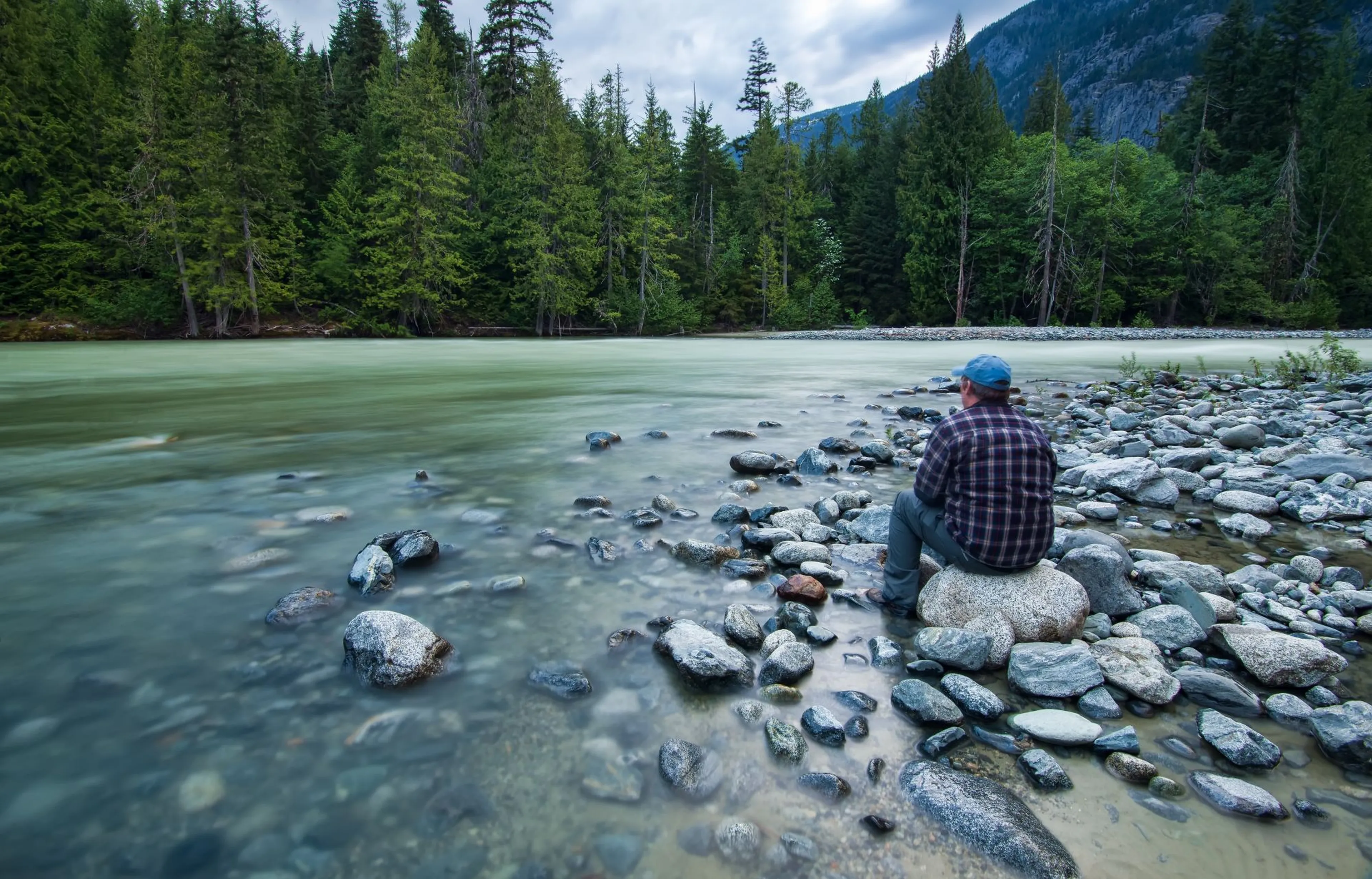 Person sitting on rocks by a gently flowing river, surrounded by forest and mountains under a cloudy sky.