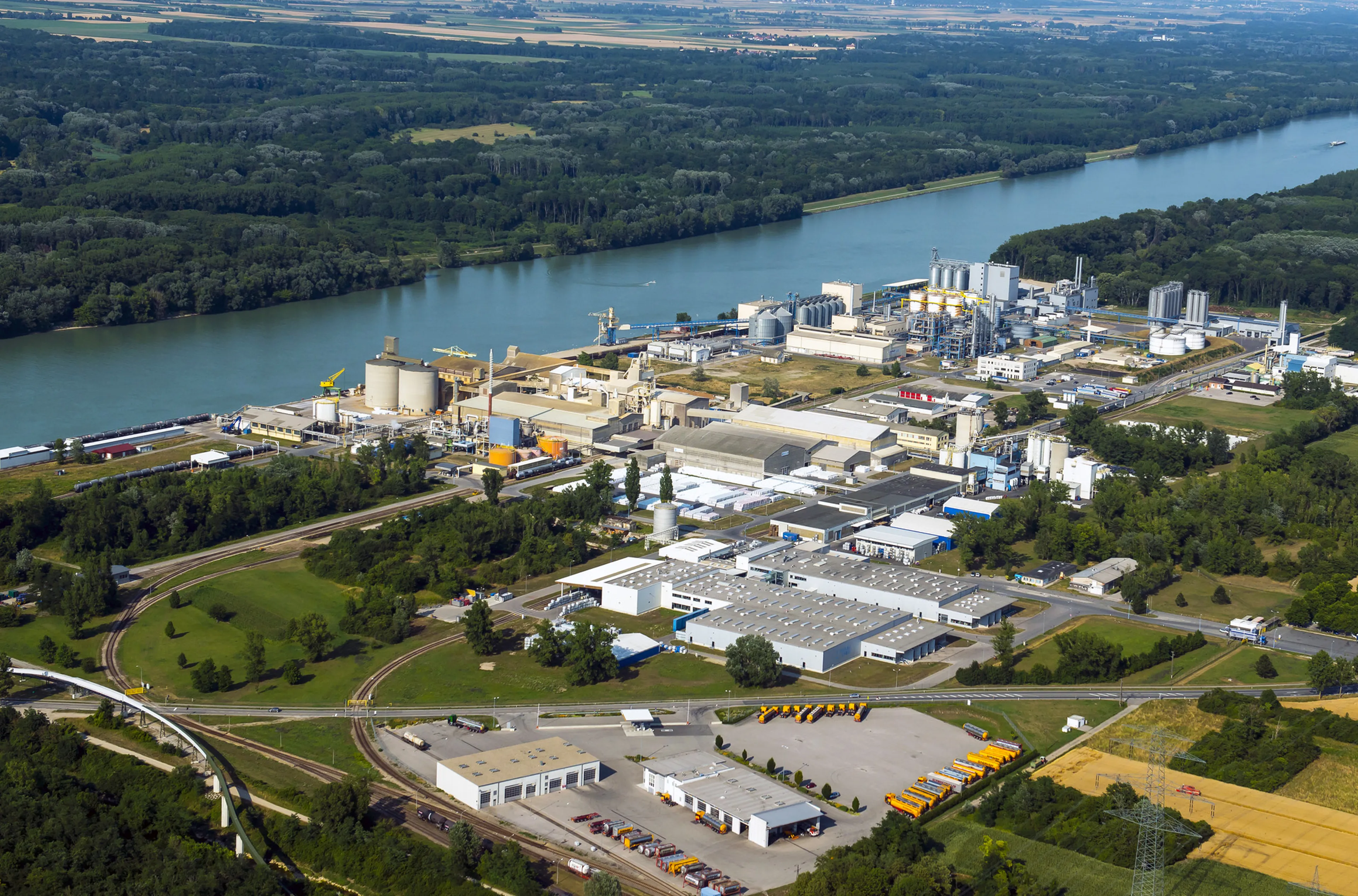 Aerial view of an industrial complex near a river, surrounded by lush greenery and fields, with various buildings and a railway track.