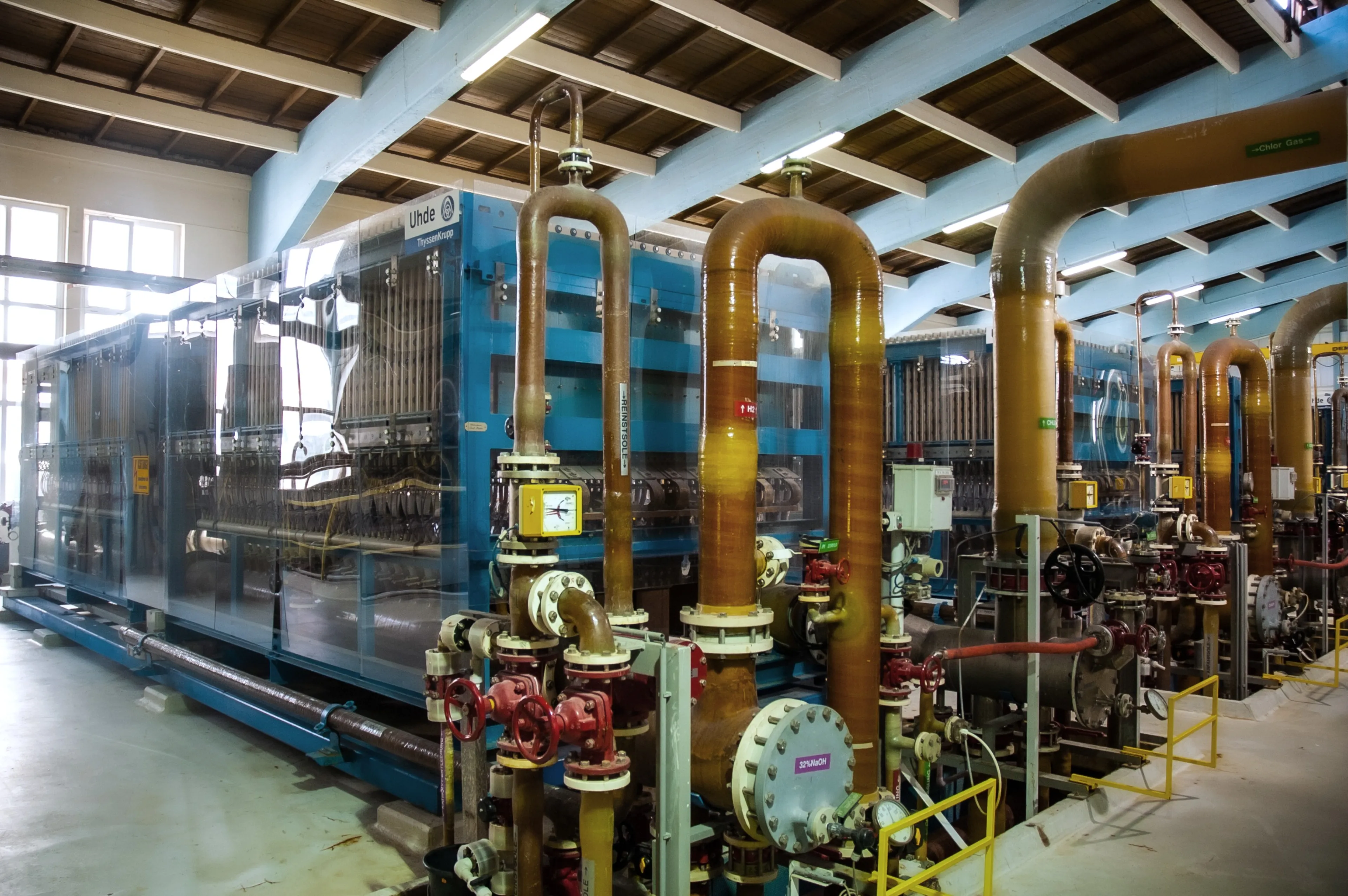 Industrial machinery with large orange pipes and blue frames inside a spacious facility, under a wooden ceiling with bright windows.