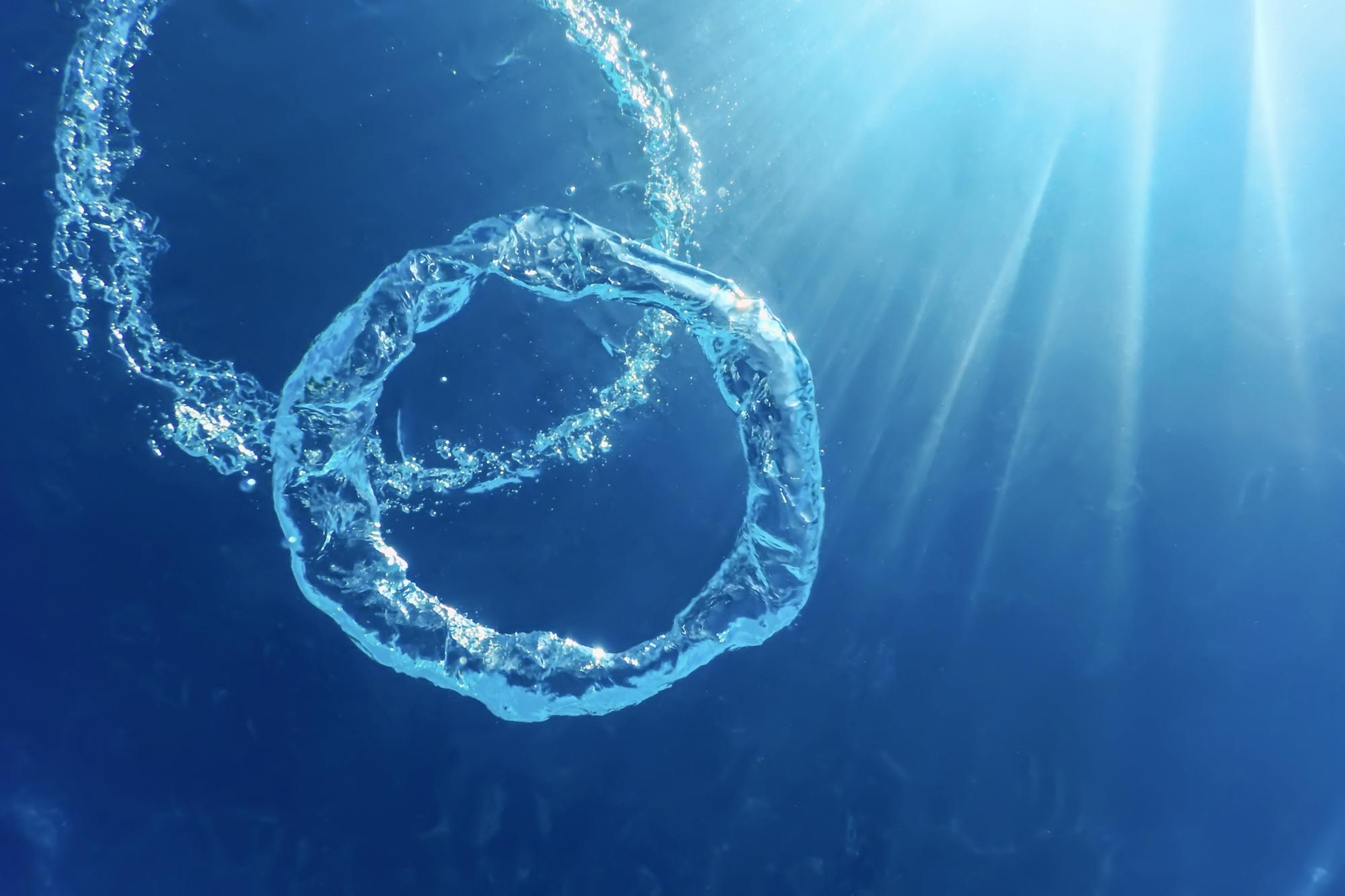 Underwater view of a clear water ring bubble ascending towards the surface, with sunlight rays filtering through the deep blue water.