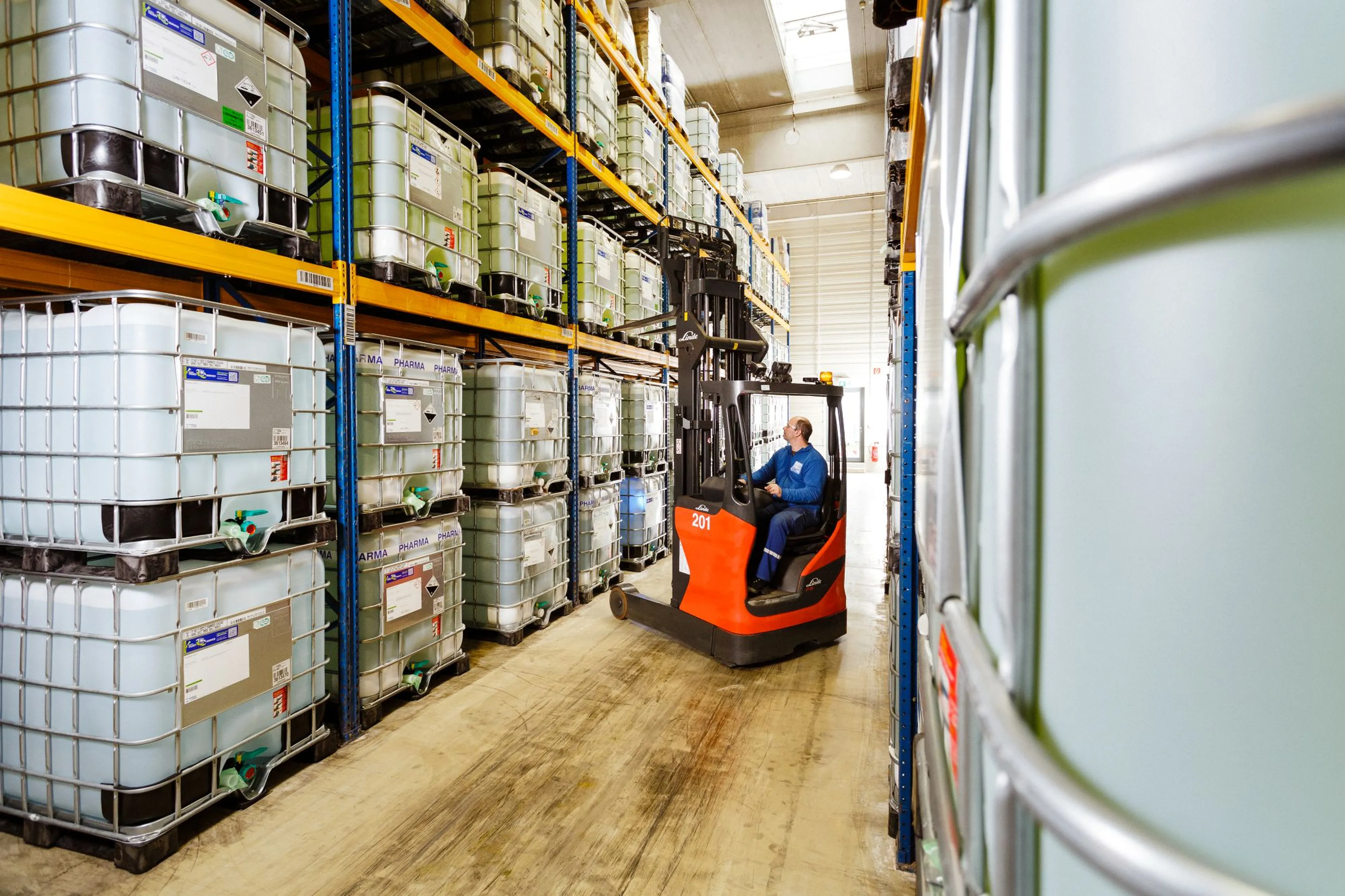 A worker operates a forklift in a warehouse, maneuvering between stacked shelves filled with large, translucent storage containers.