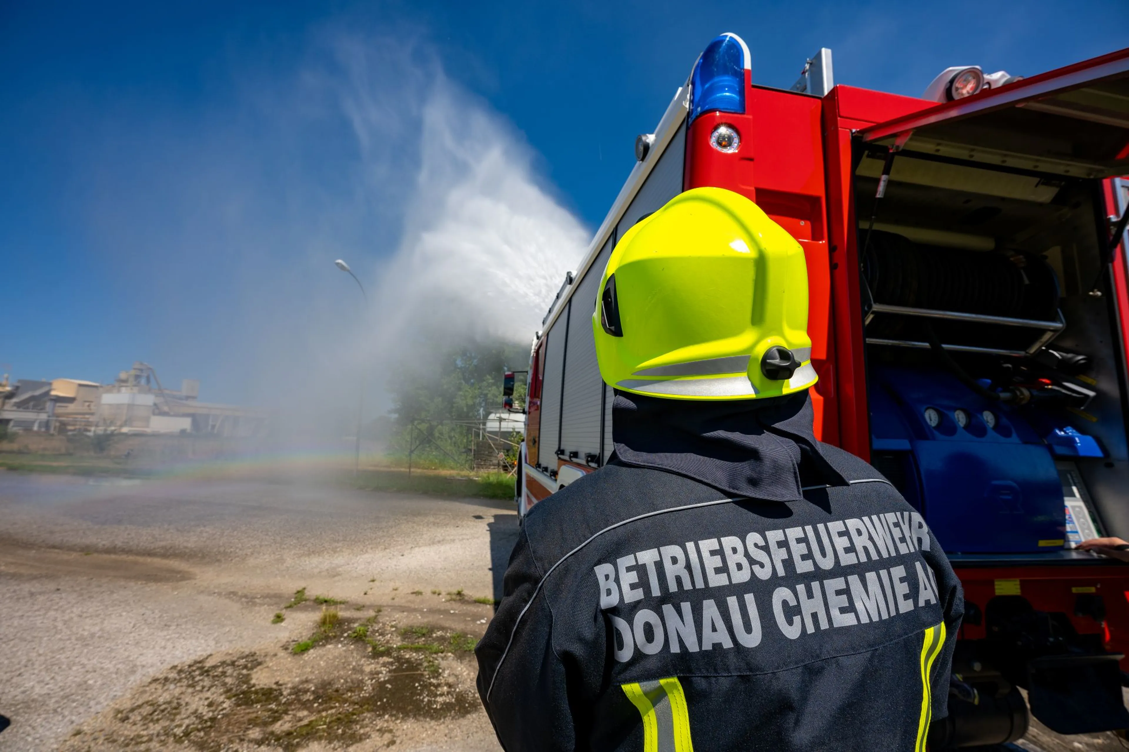 A firefighter in a neon helmet sprays water from a fire truck hose, creating a rainbow. The truck is marked "Betriebsfeuerwehr Donau Chemie AG."