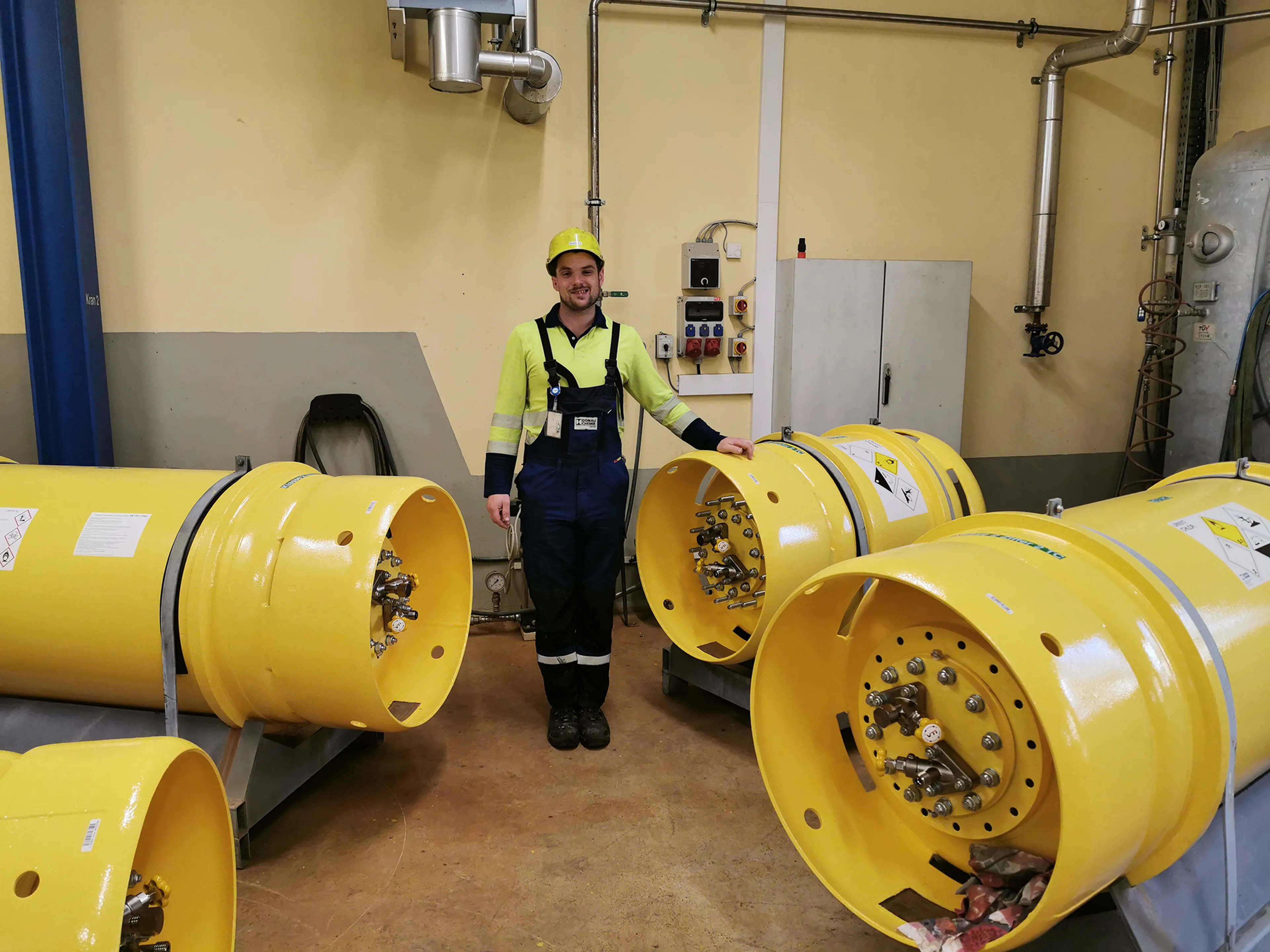 A worker in a high-visibility outfit and helmet stands beside large yellow industrial equipment in a workshop.