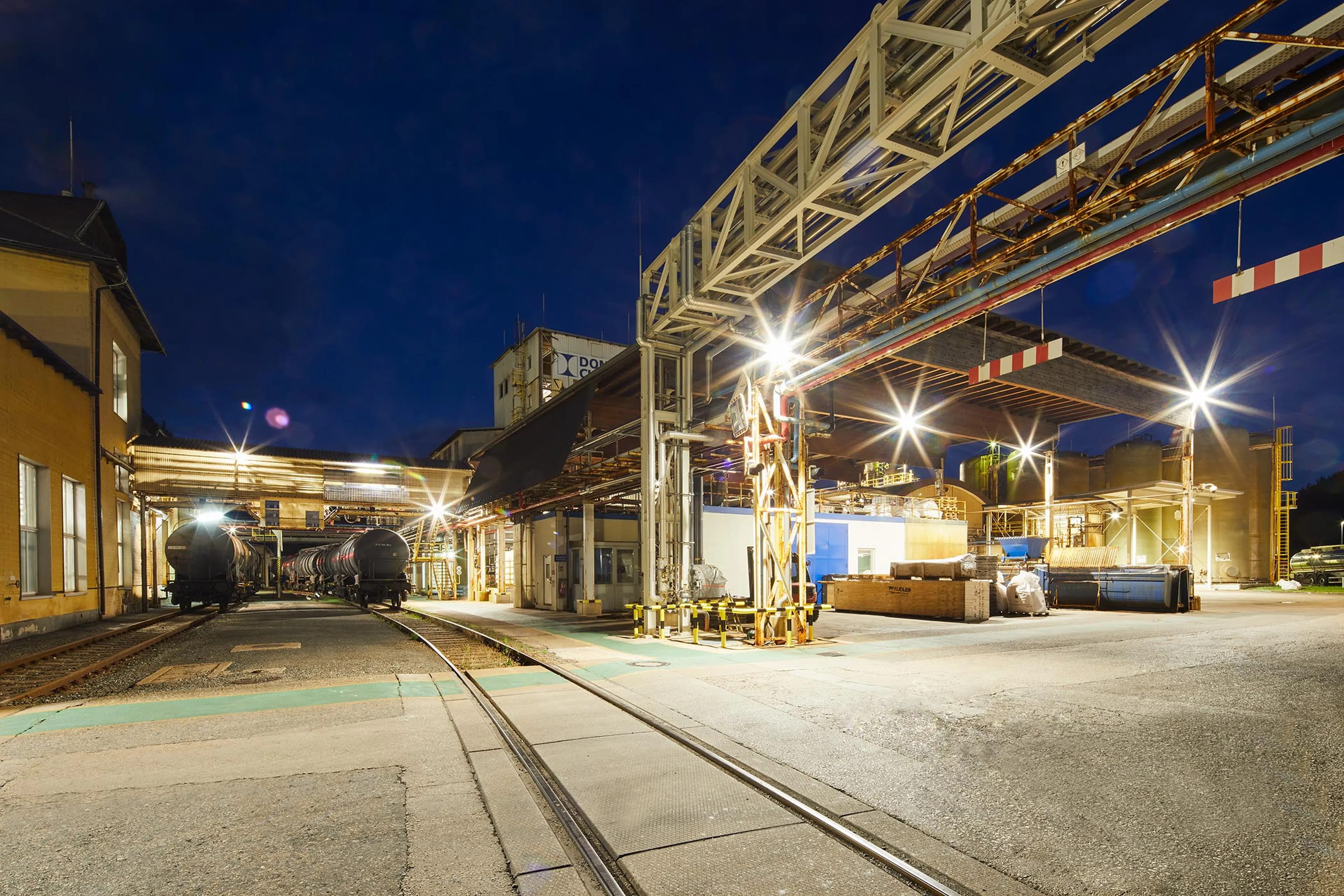 Industrial train station at night with bright lights, tracks, overhead structures, and buildings in the background under a dark blue sky.