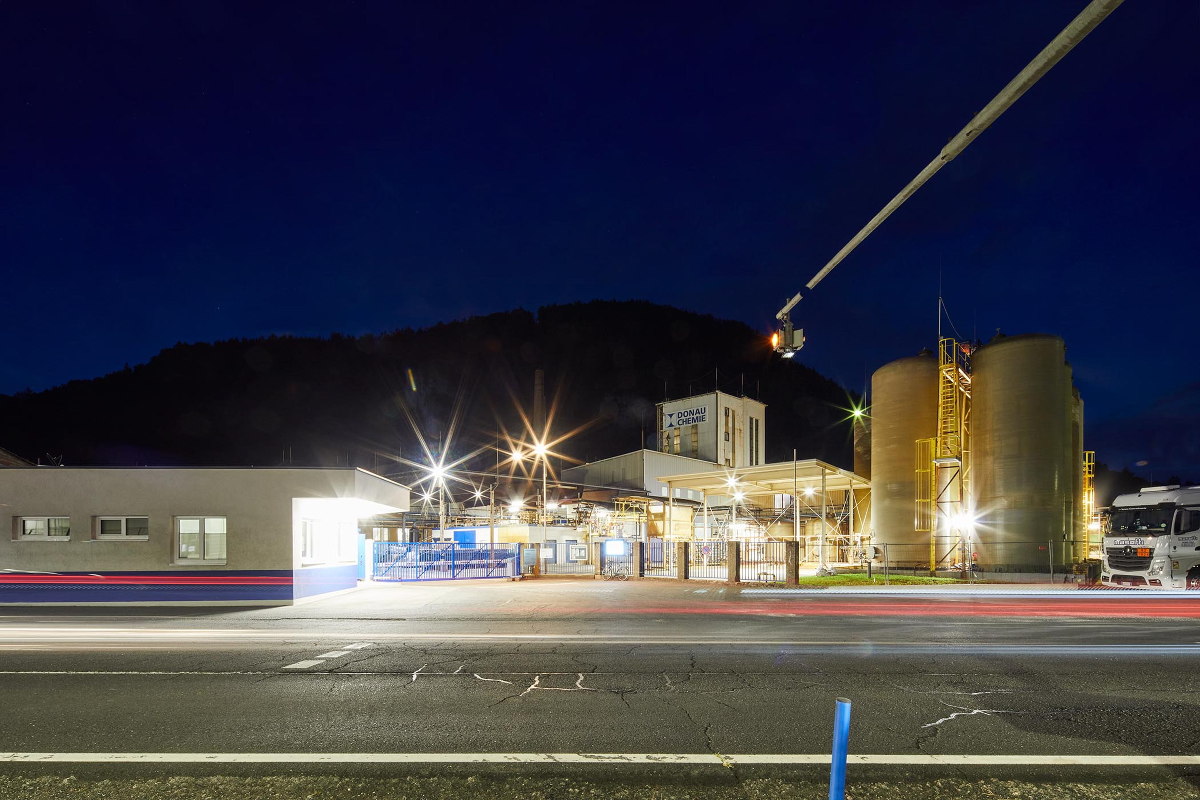 Industrial facility at night with illuminated tanks and buildings, surrounding mountains, and blurred lines from passing vehicle lights.