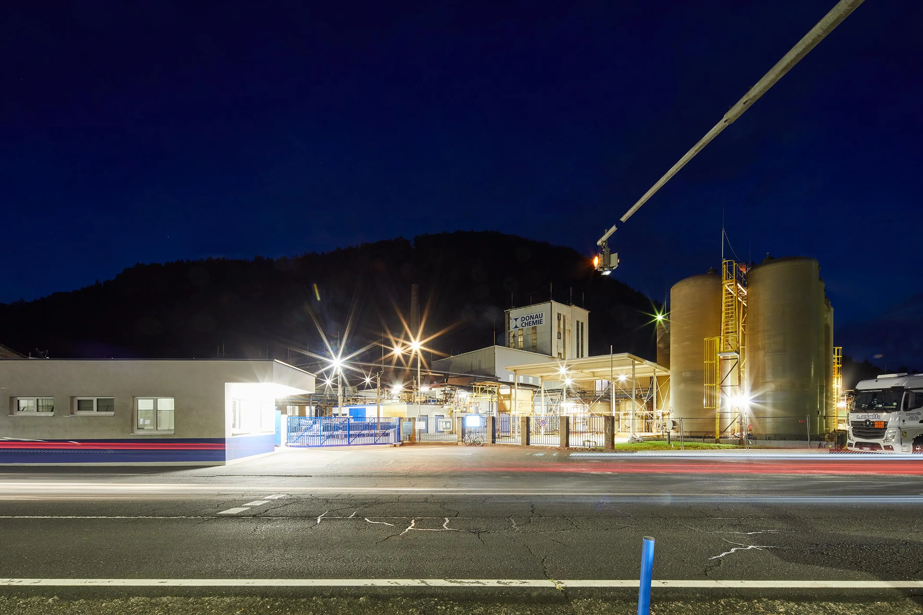 Industrial facility at night with illuminated tanks and buildings, surrounding mountains, and blurred lines from passing vehicle lights.