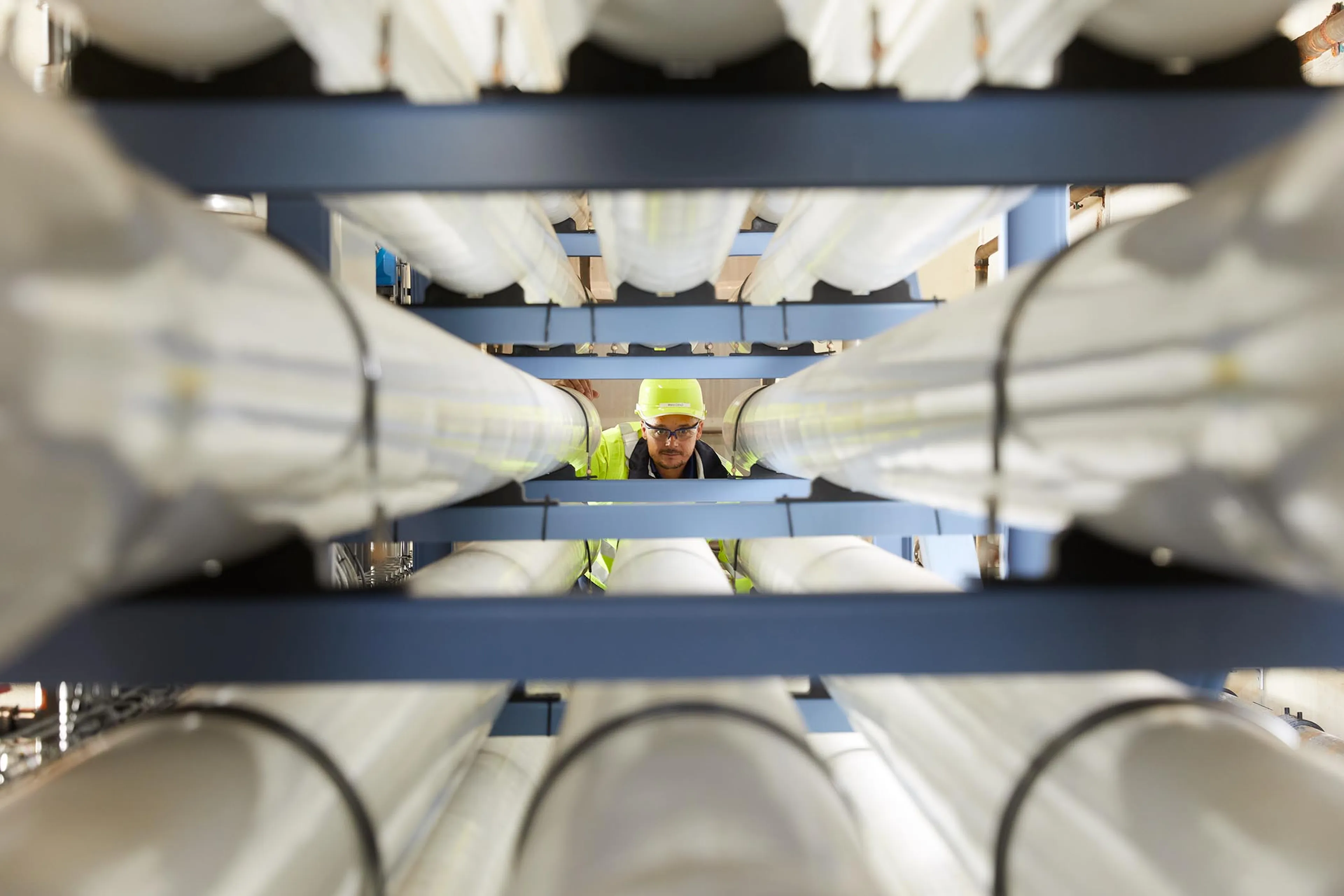 Worker in a yellow hard hat inspecting large cylindrical pipes arranged in parallel, viewed from between the pipes.