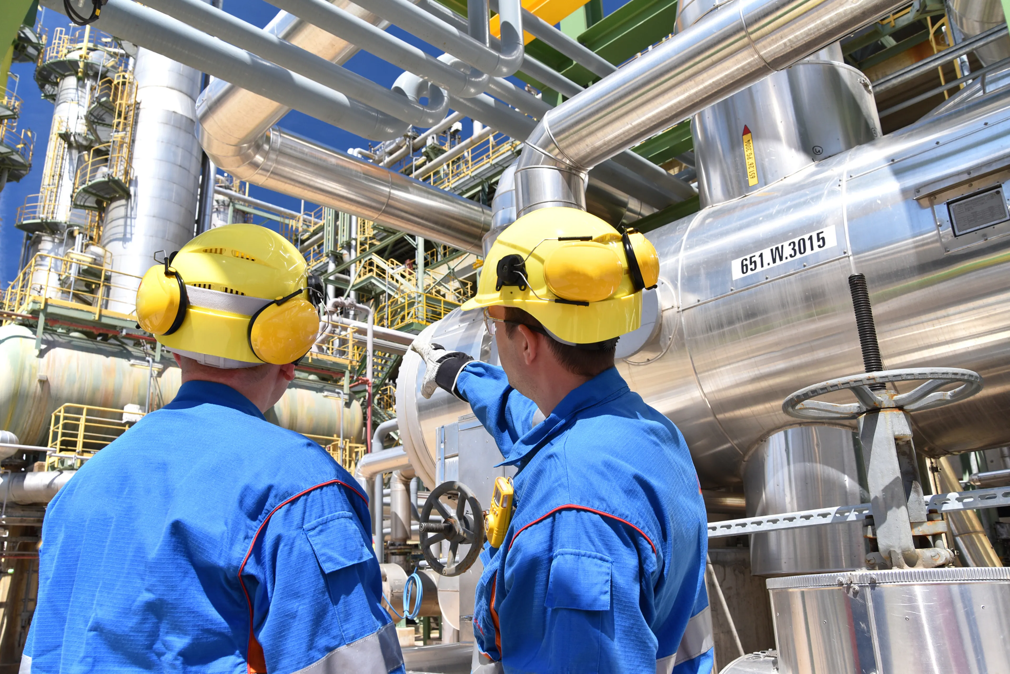 Two workers in blue protective gear and yellow helmets discuss in a complex industrial setting with metal pipes and equipment.