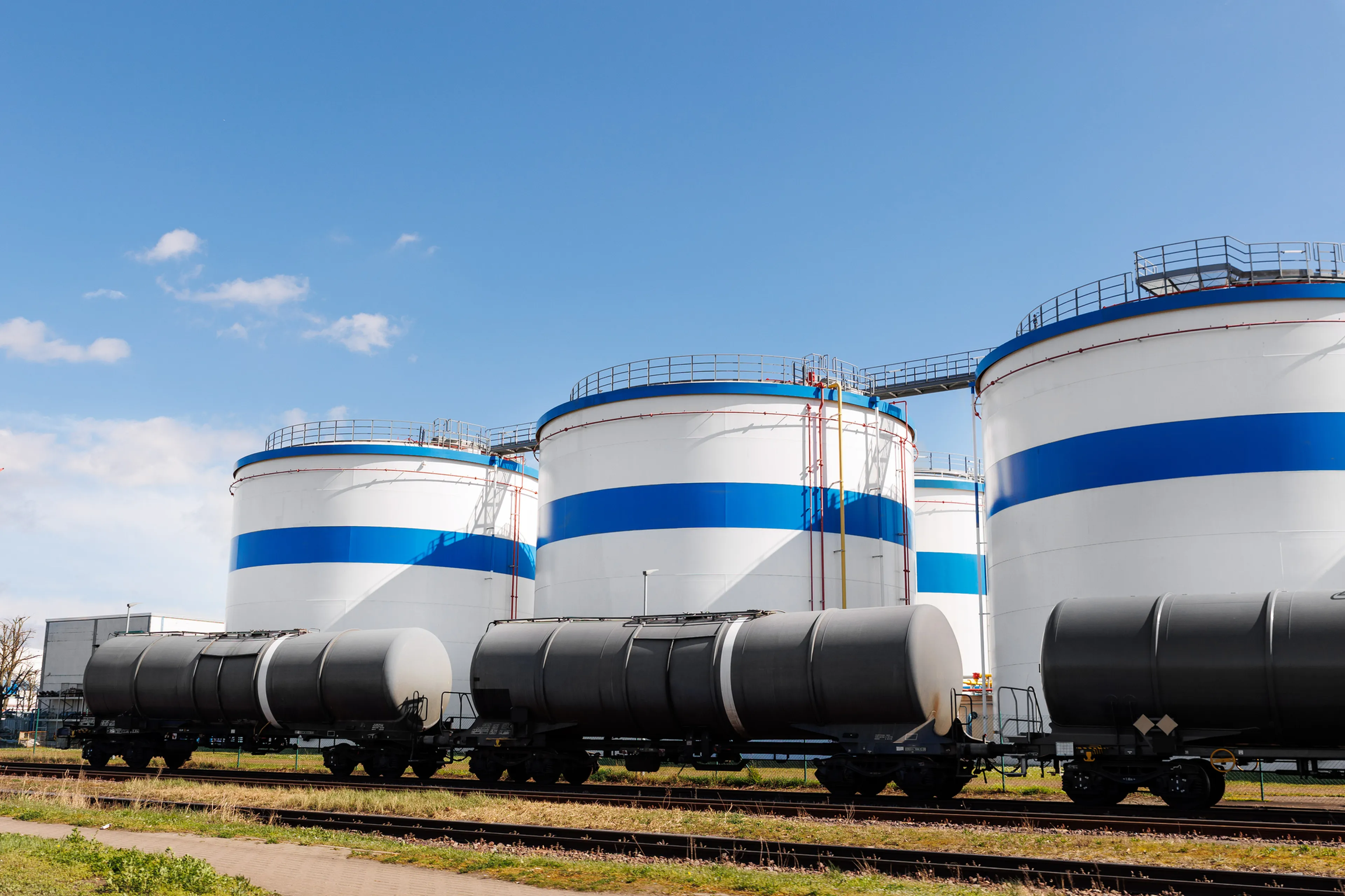 Large industrial storage tanks with blue stripes, adjacent to railway tracks and rail tank cars, under a clear blue sky.